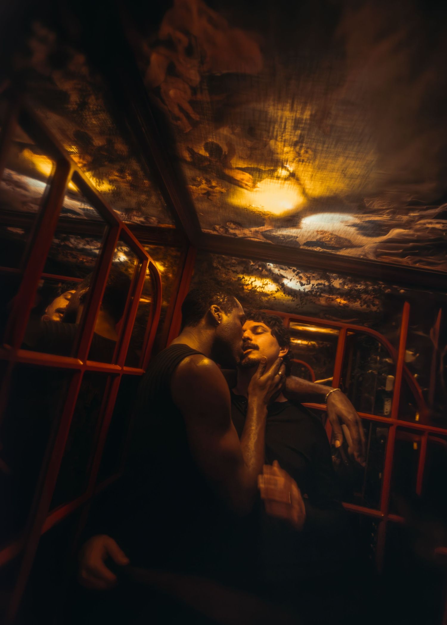 Two figures in dramatic warm light inside a dark, moody interior with red metal bars and golden ceiling.