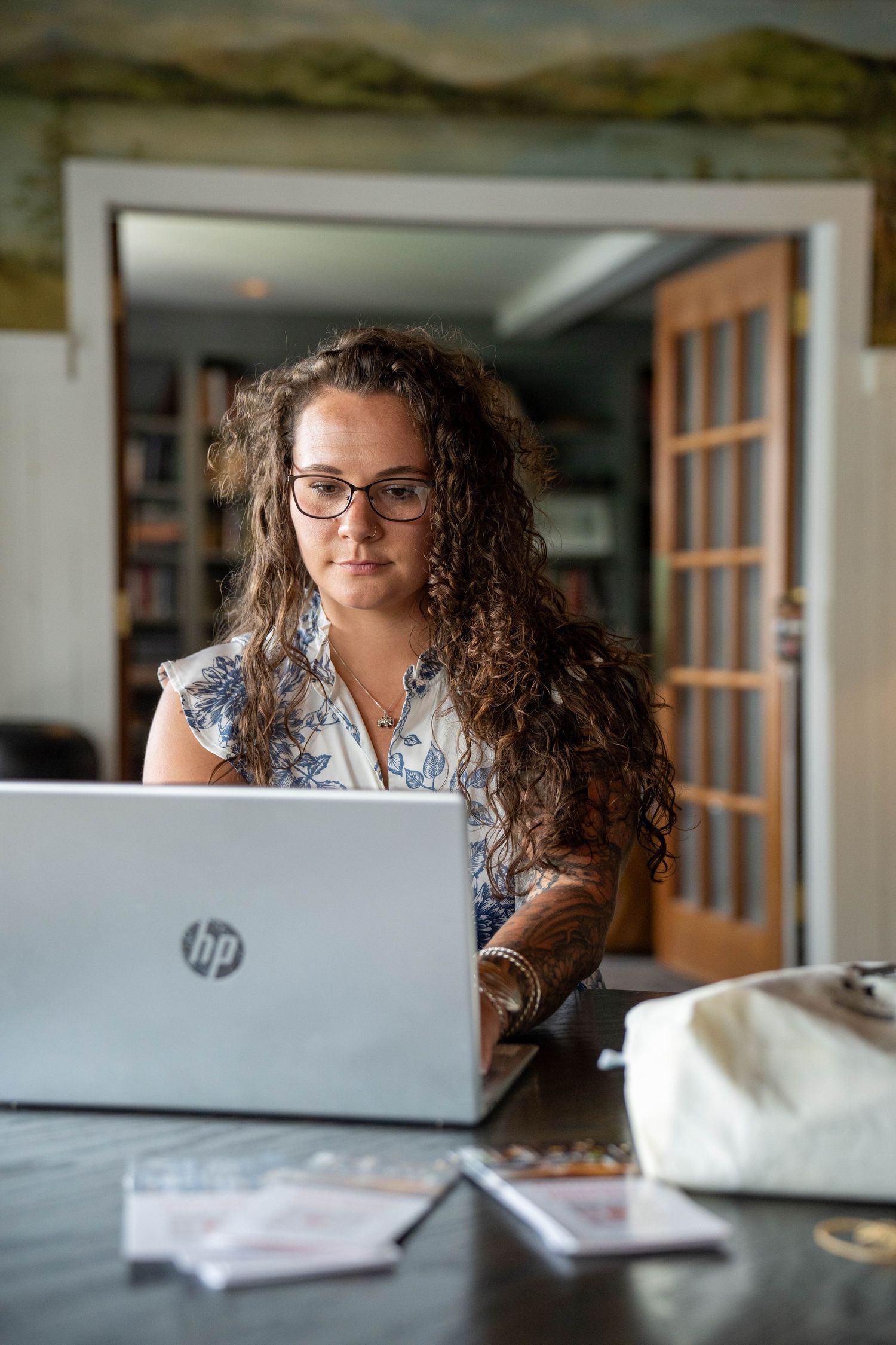 Woman with curly hair and glasses working on an HP laptop at a desk in a home office setting.