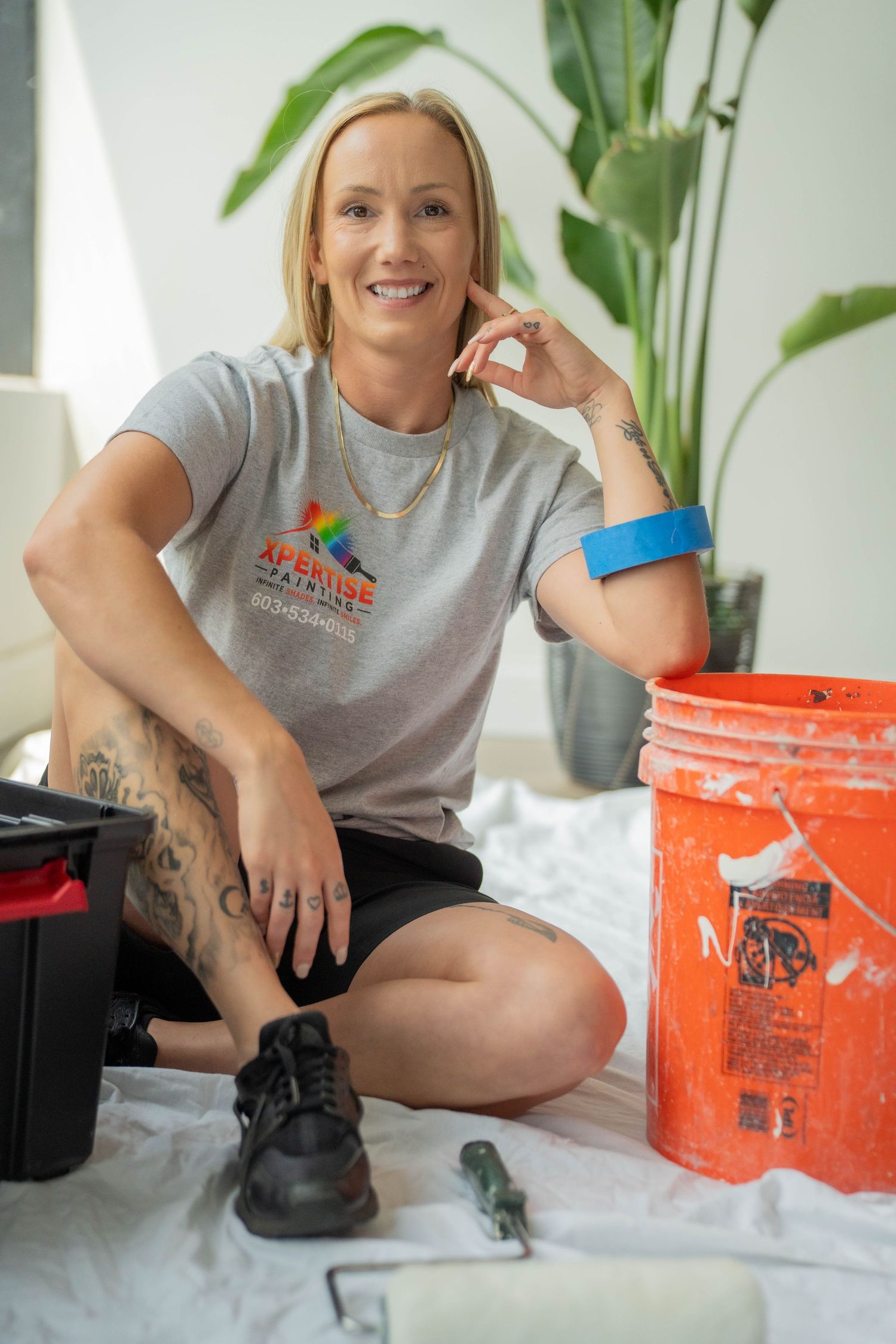 A smiling female painter sits on a drop cloth with a red paint bucket and tools, wearing a gray Aprise Painting t-shirt.