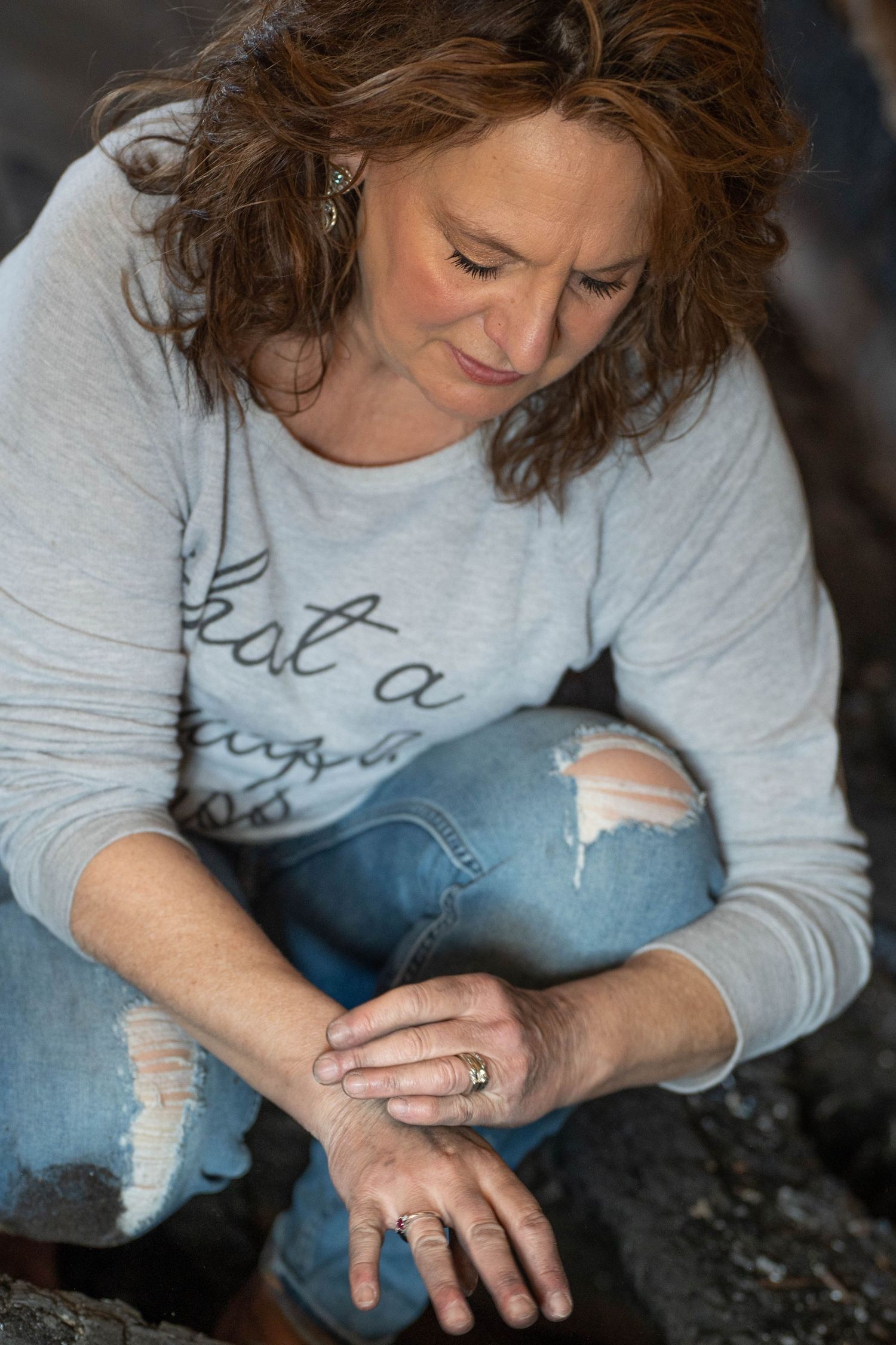 A middle-aged woman with curly auburn hair sits on rocks, wearing ripped jeans and a gray sweatshirt, looking down at her hands.