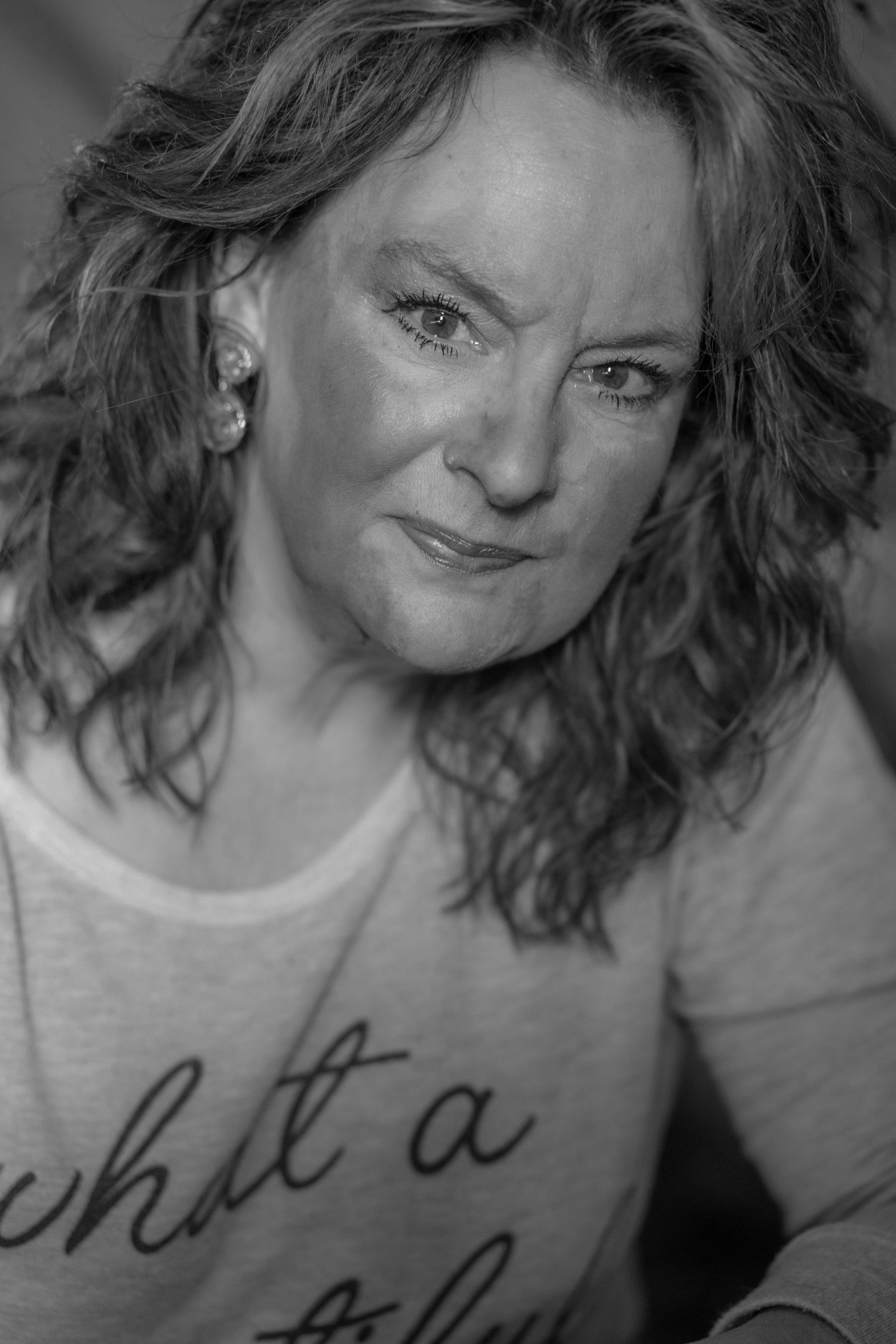 Black and white portrait of a middle-aged woman with curly hair wearing a graphic tee, smiling softly at the camera.