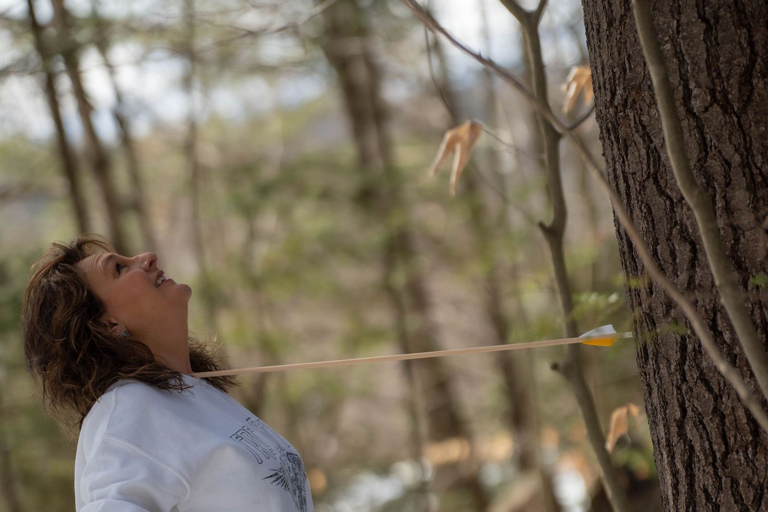 Woman in white jacket looks up at an arrow flying past a tree in a forest setting.