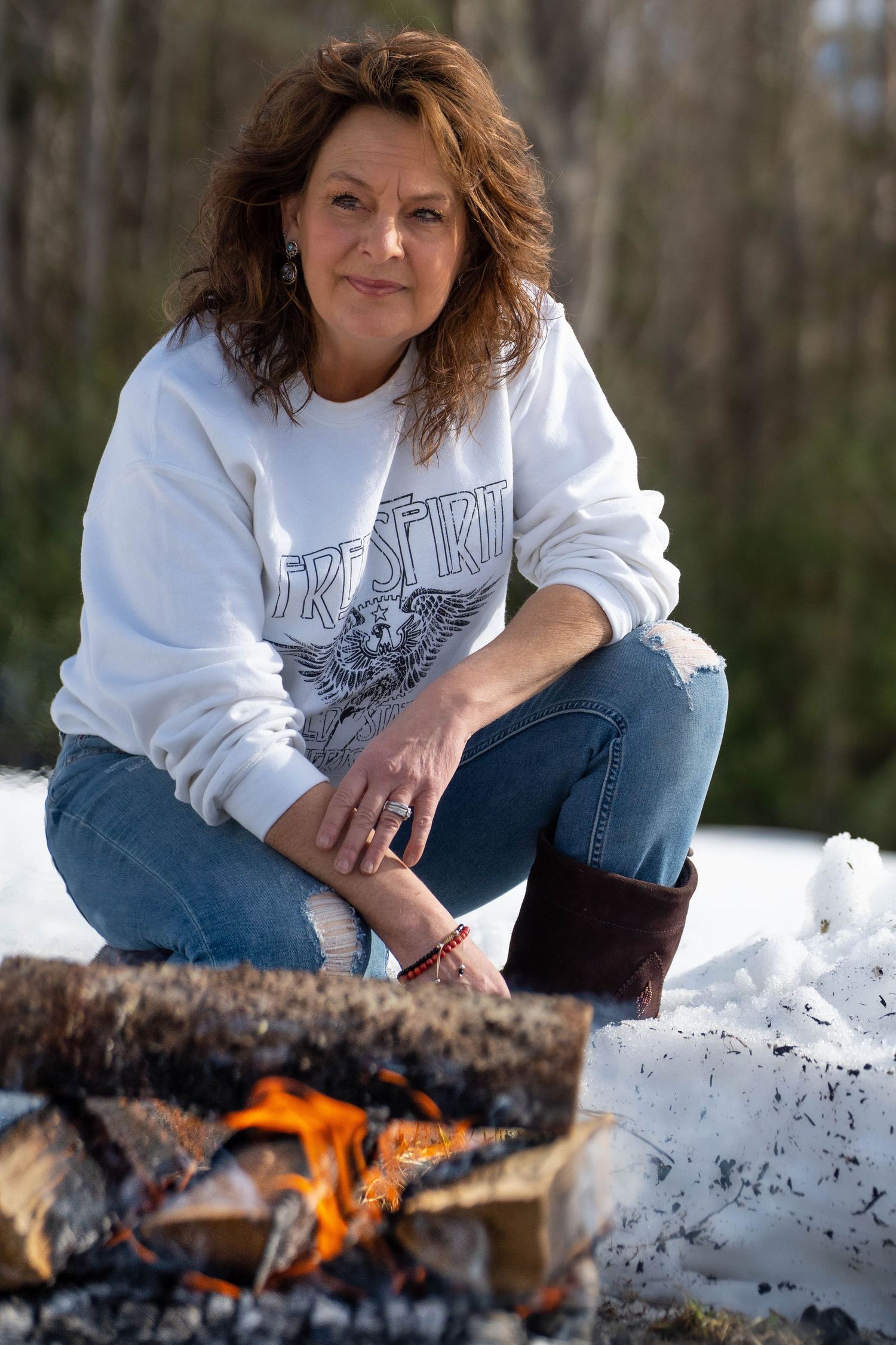 Woman in white sweatshirt and jeans crouching by a campfire in the snow, warming her hands outdoors.