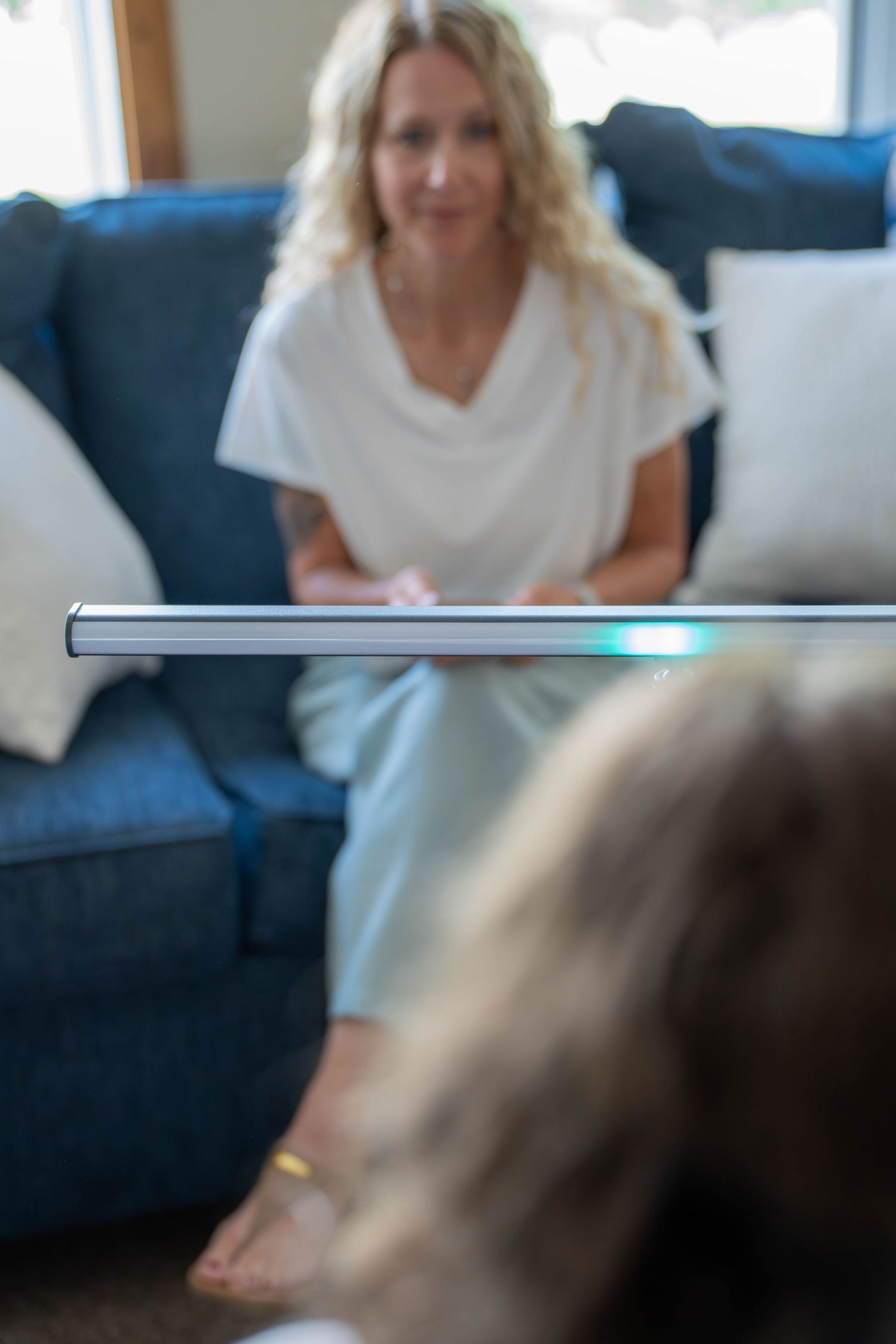 Blonde woman in white outfit sits on blue sofa during therapy session, with EMDR light bar in foreground.