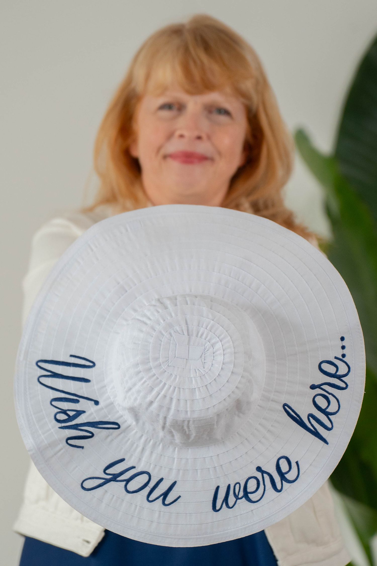 Woman holding a wide-brim white sun hat with "Wish you were here..." written in navy blue script.