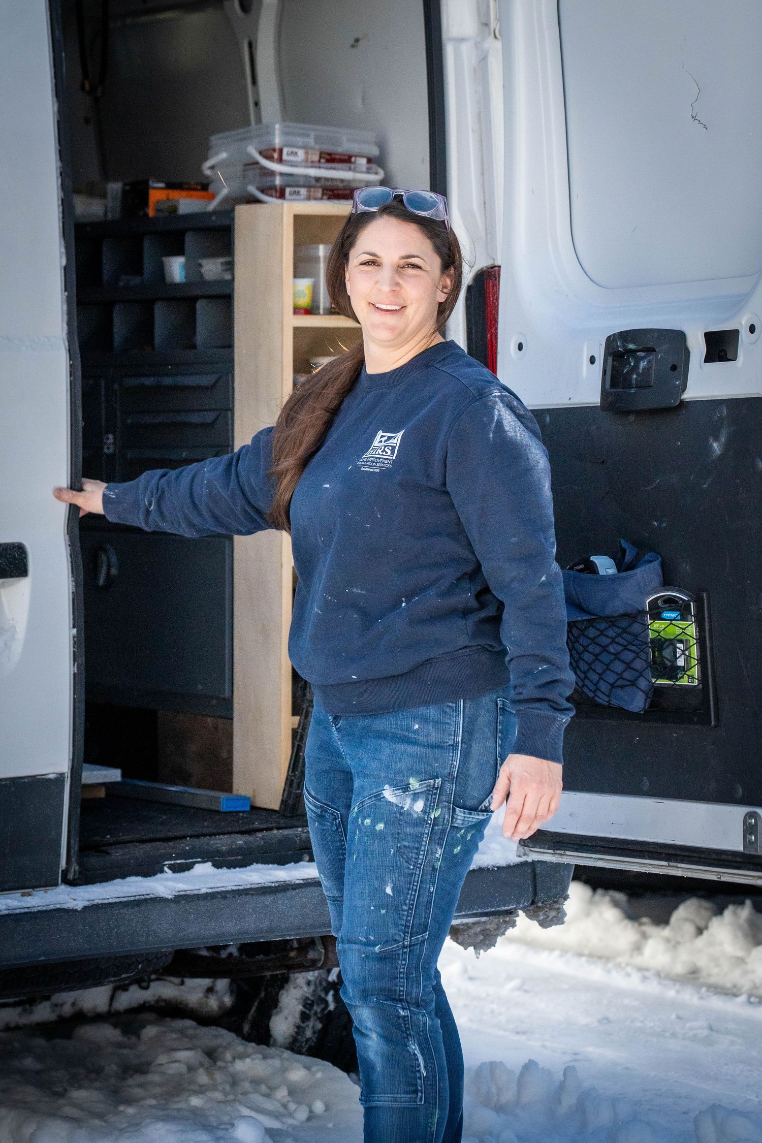 Woman in navy sweatshirt and jeans smiles beside open cargo van doors in snowy conditions.