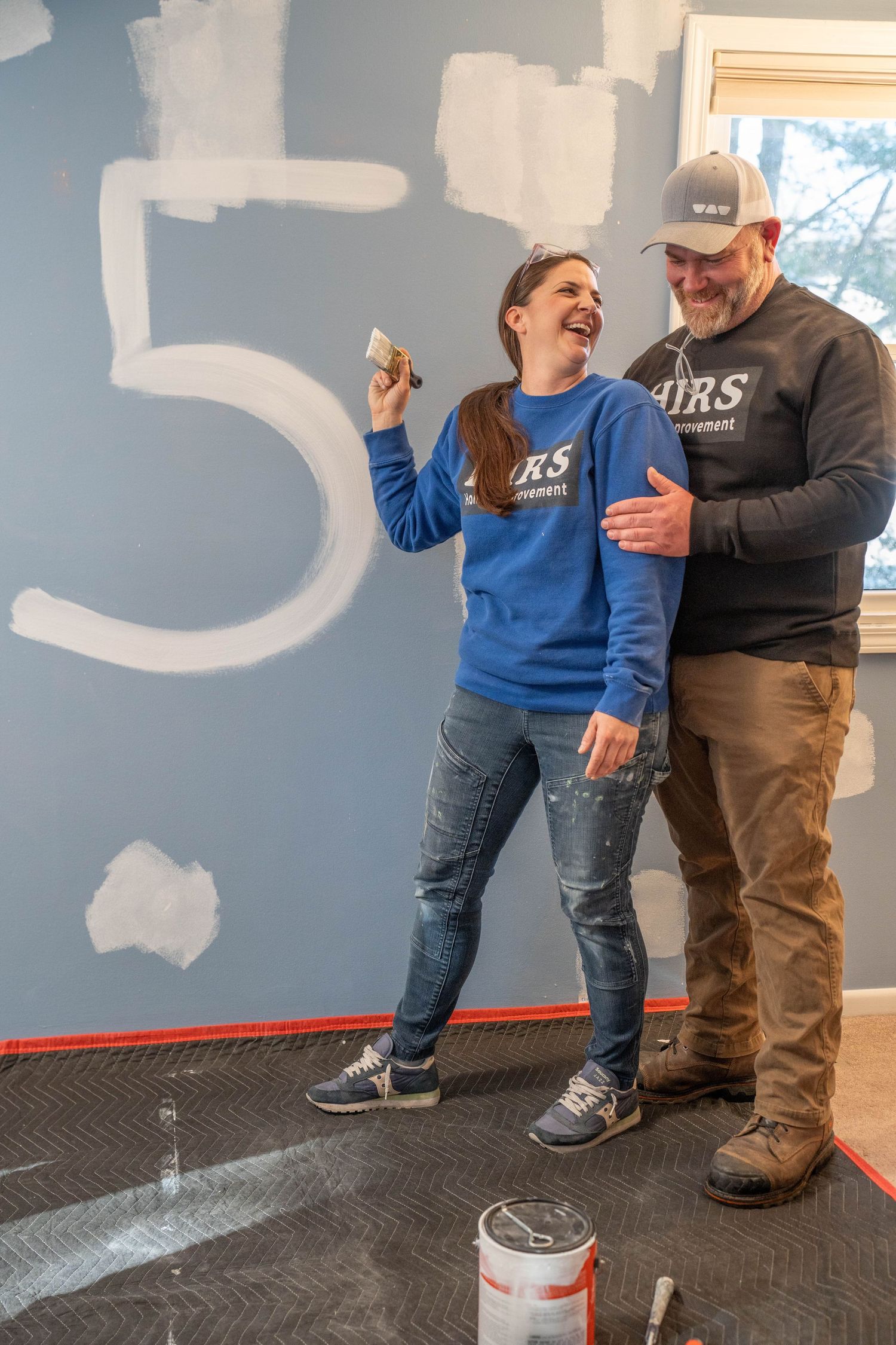 A smiling couple in matching branded sweatshirts painting a large number 5 on a blue wall indoors.