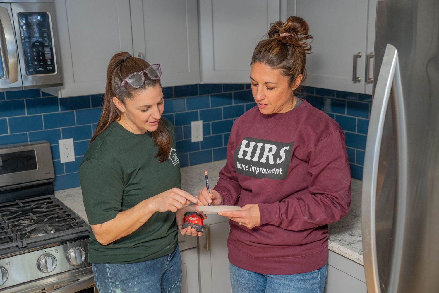 Two women reviewing home improvement plans in a modern kitchen with stainless steel appliances and blue tile backsplash.