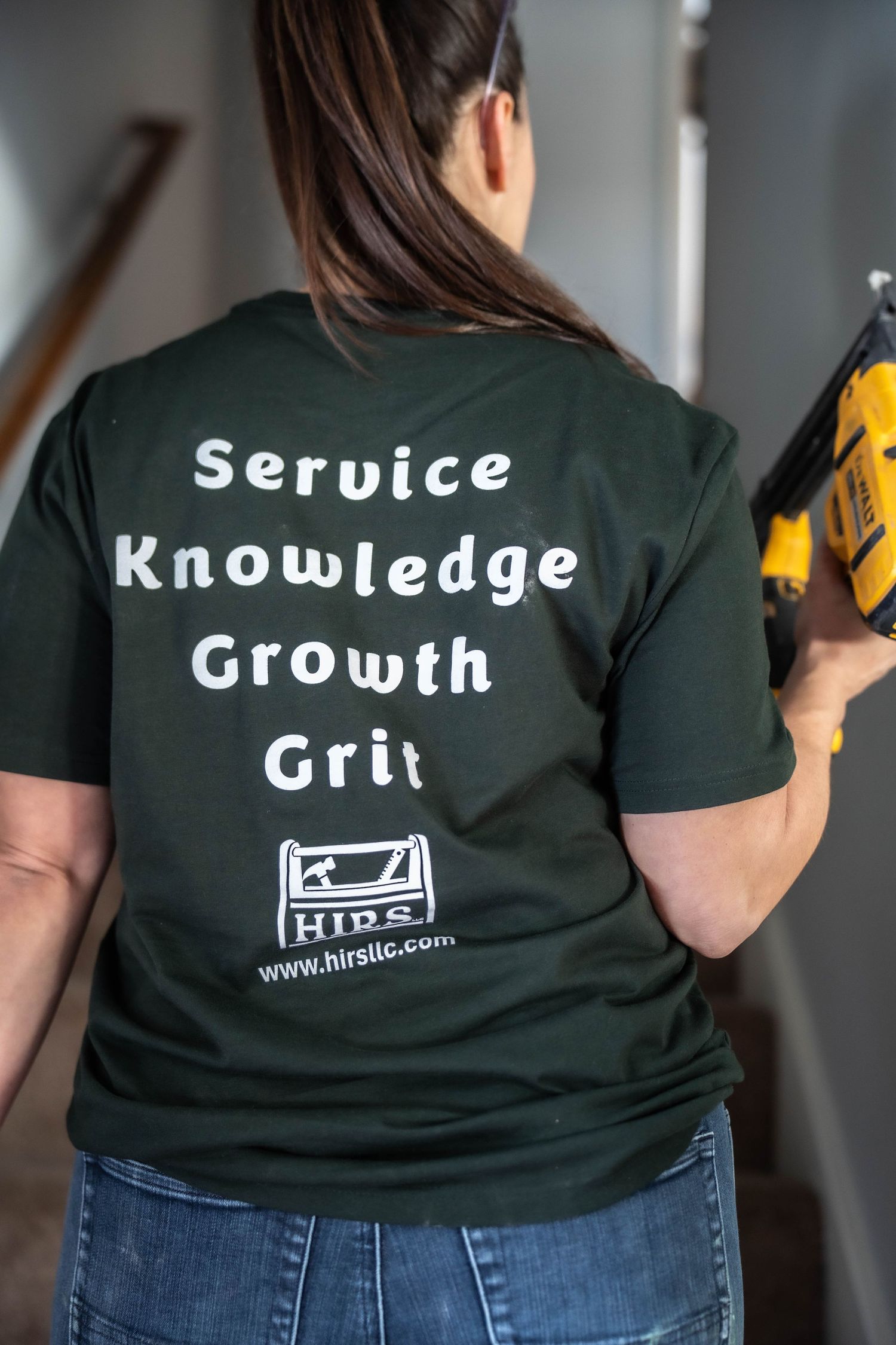 Woman wearing dark green HIRS LLC t-shirt reading Service Knowledge Growth Grit, holding a power nail gun.