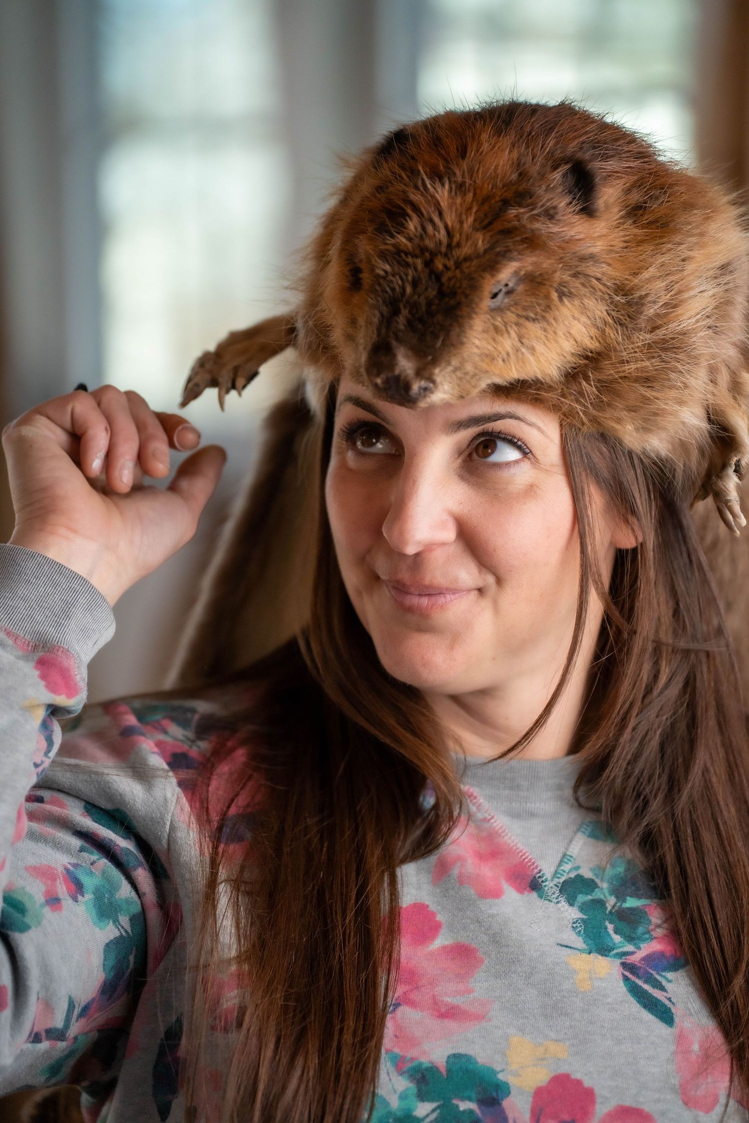 A young woman wearing a fur animal pelt hat holds it on her head while smiling indoors.