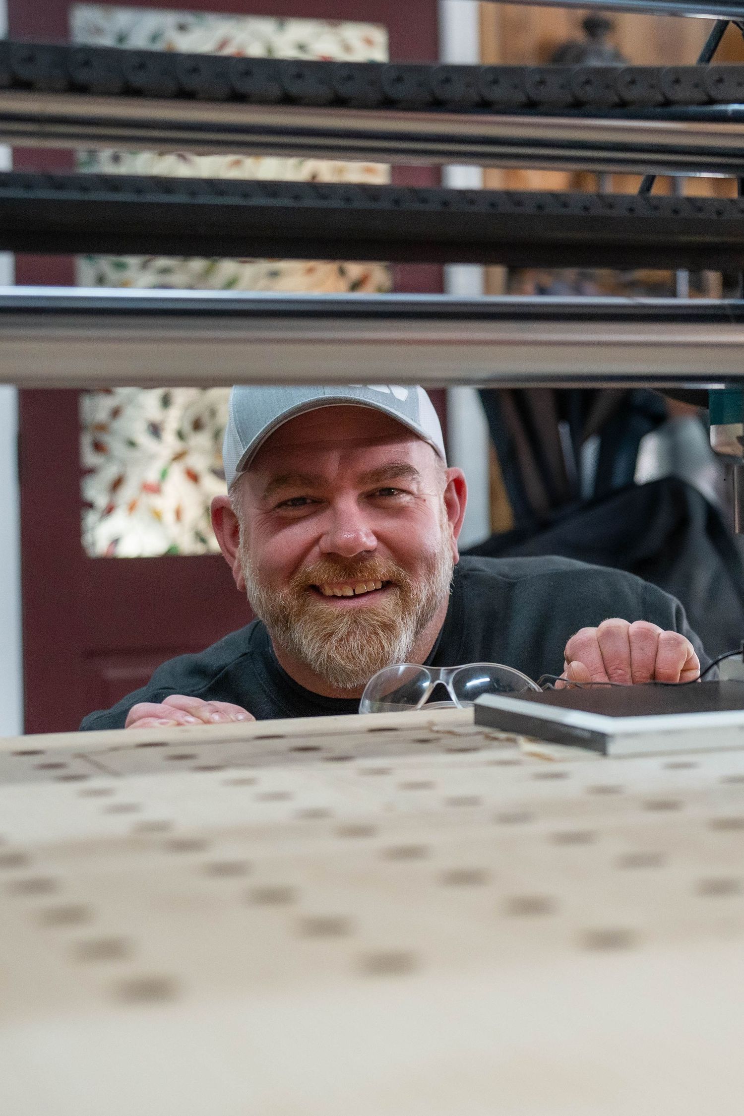 Smiling bearded man in gray cap peers over a wooden workbench with glasses and tools in a workshop.