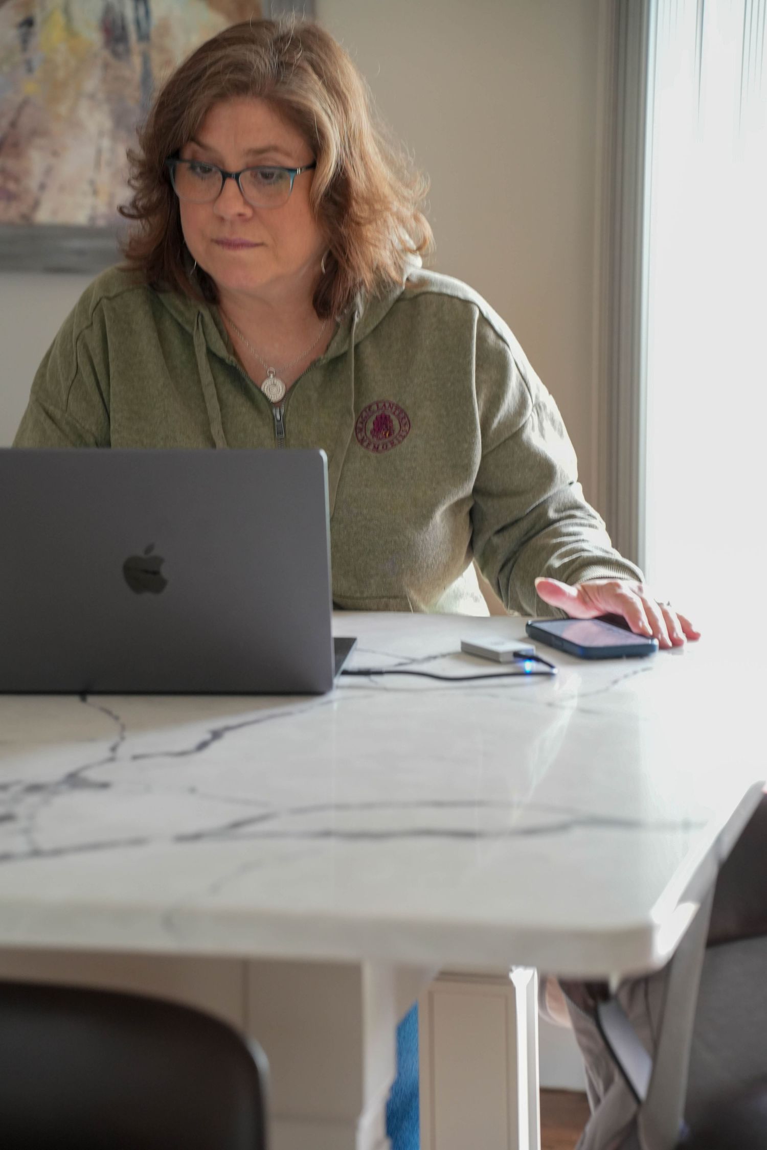 Woman in green hoodie working on Apple MacBook laptop at marble table with smartphone nearby.