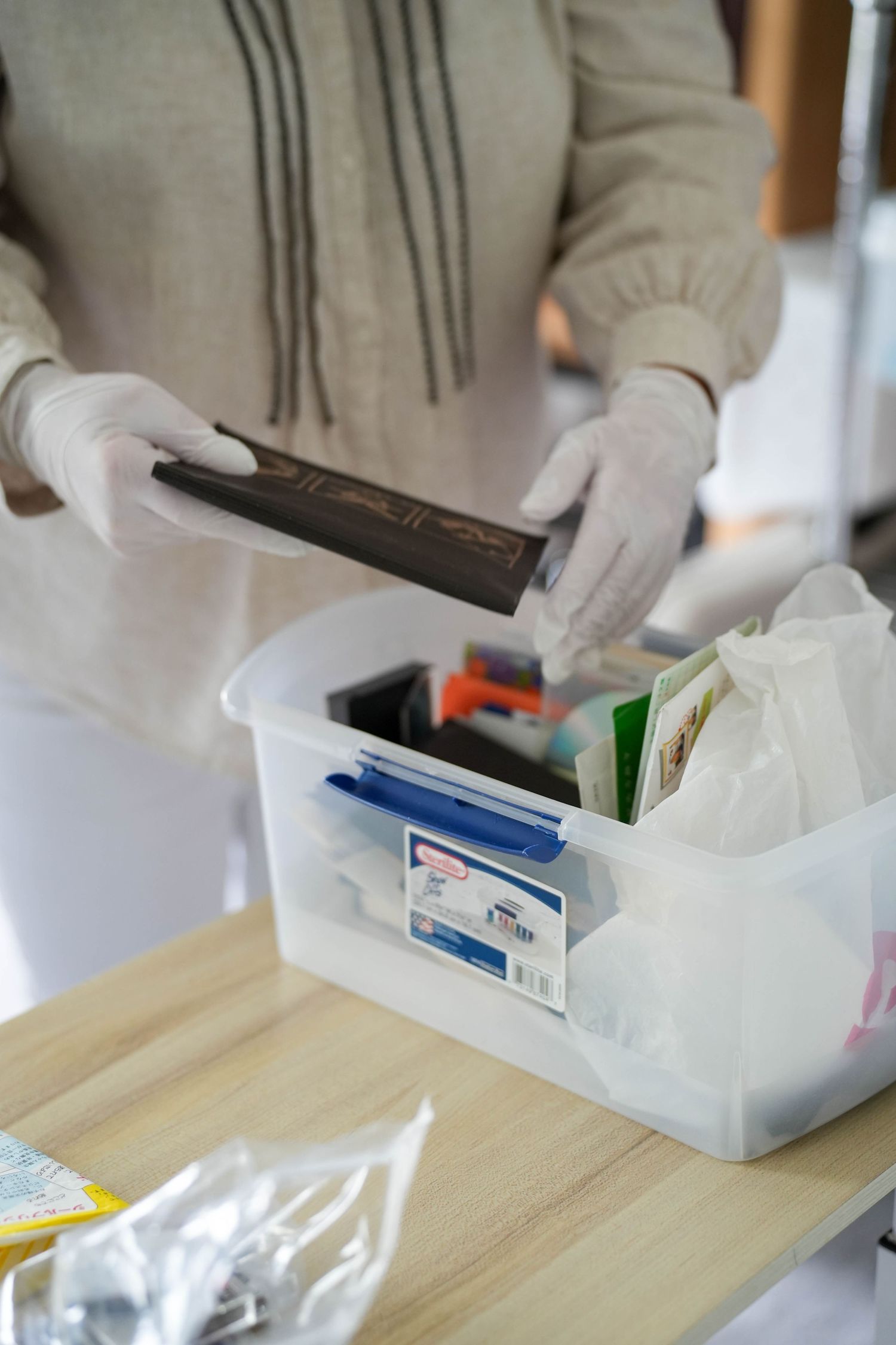 Person wearing latex gloves sorting items into a clear plastic storage container on a wooden table.