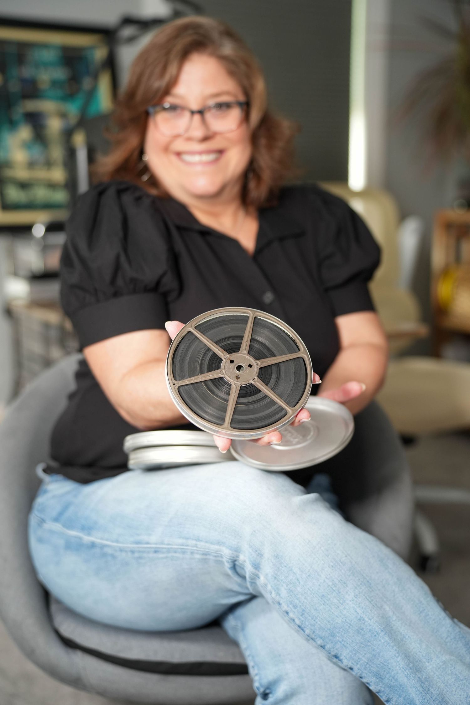 Smiling woman in black shirt and jeans sitting in chair holding vintage film reel toward camera in modern office setting.