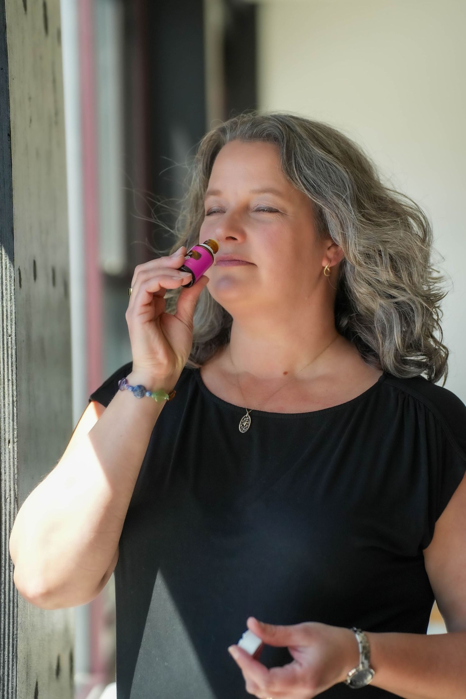Middle-aged woman with gray hair smelling essential oil from pink bottle outdoors in natural light.