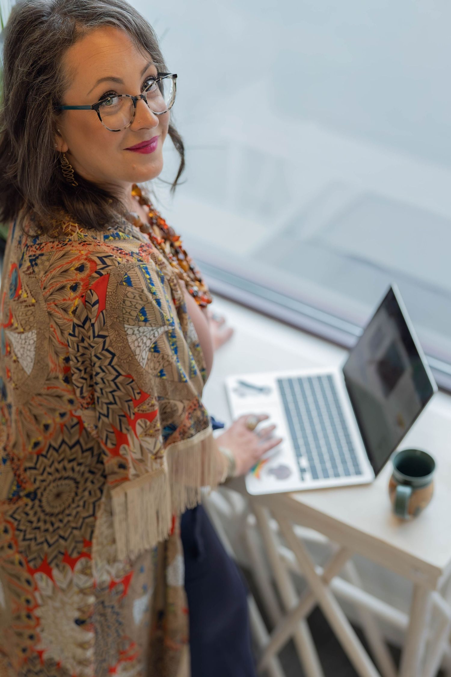 Woman in colorful patterned shawl and glasses working on laptop at standing desk, smiling at camera.