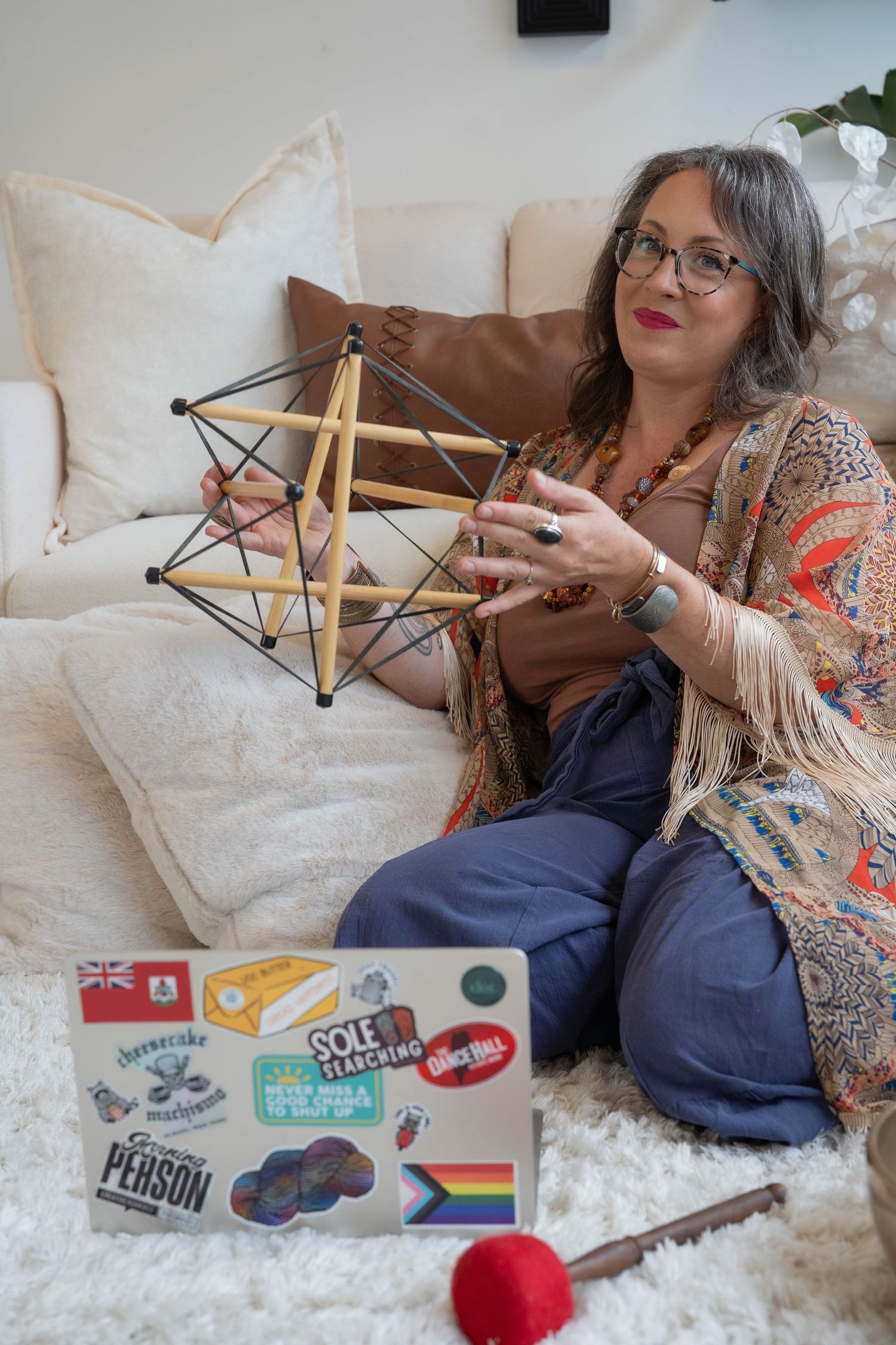 Woman sitting on white sofa holding geometric wooden star shape, with sticker-covered laptop nearby.