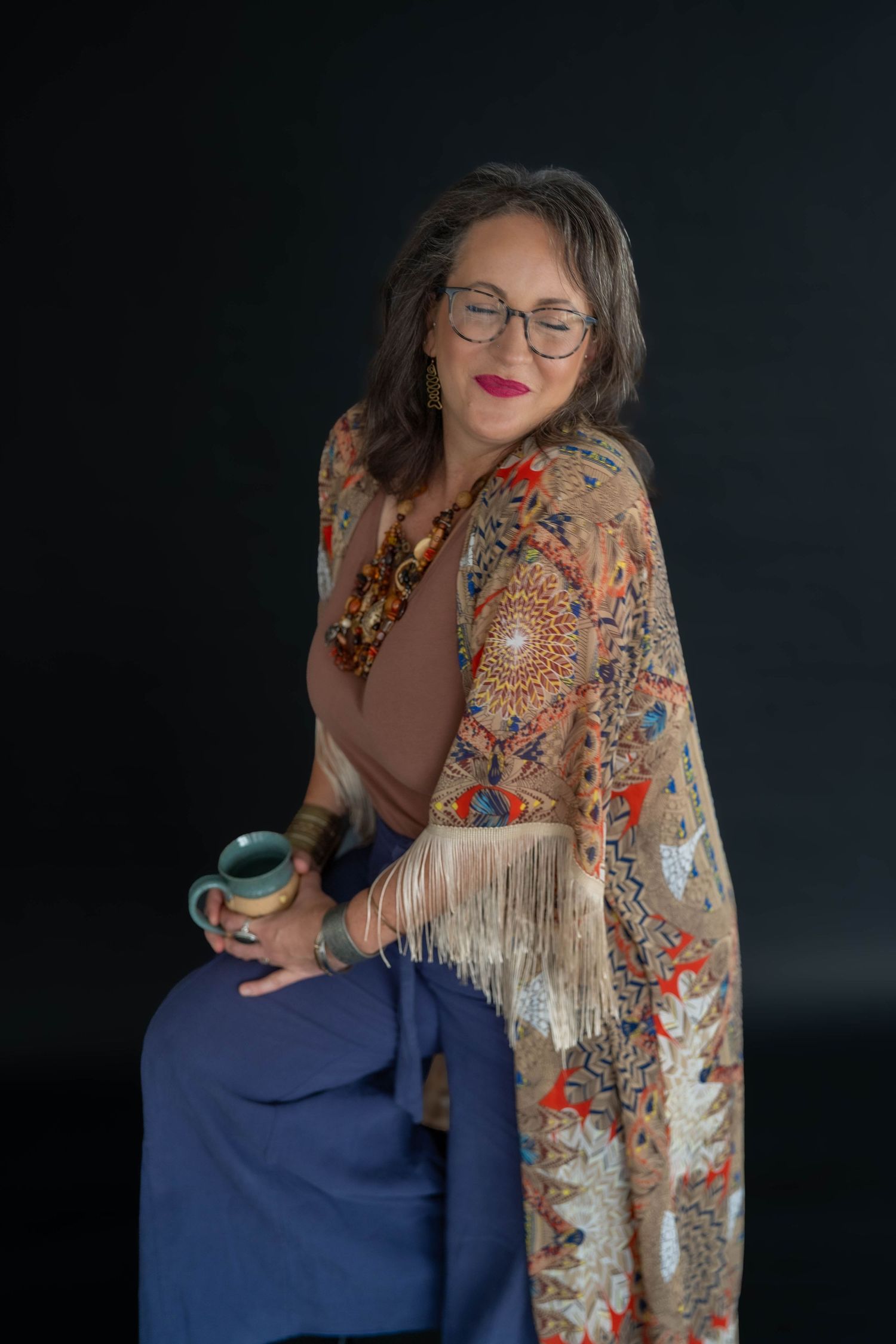 Mature woman with glasses wearing colorful fringed shawl and blue pants, holding a mug while posing against dark background.