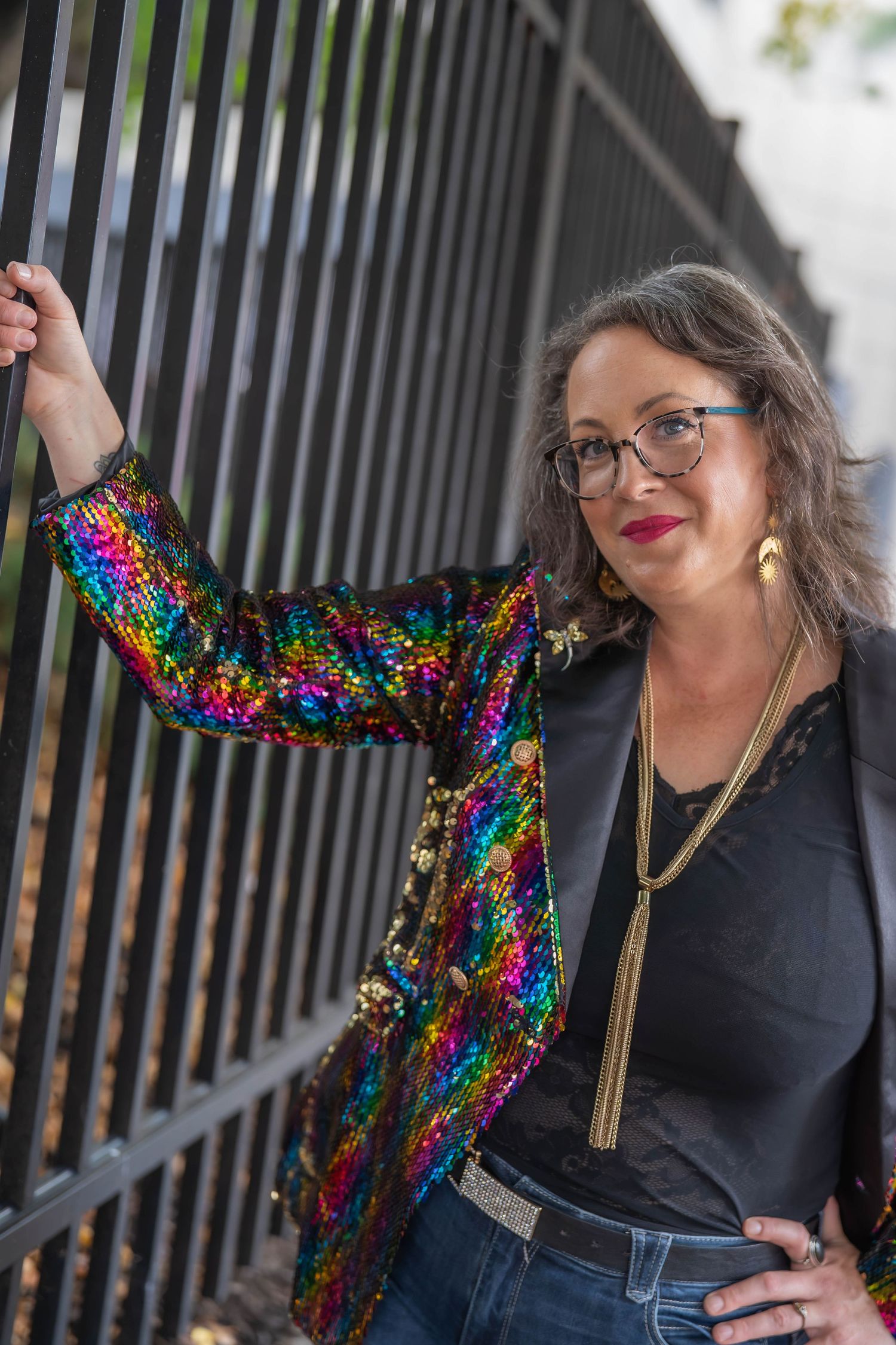 Woman in rainbow sequin blazer and glasses leaning against metal fence outdoors.