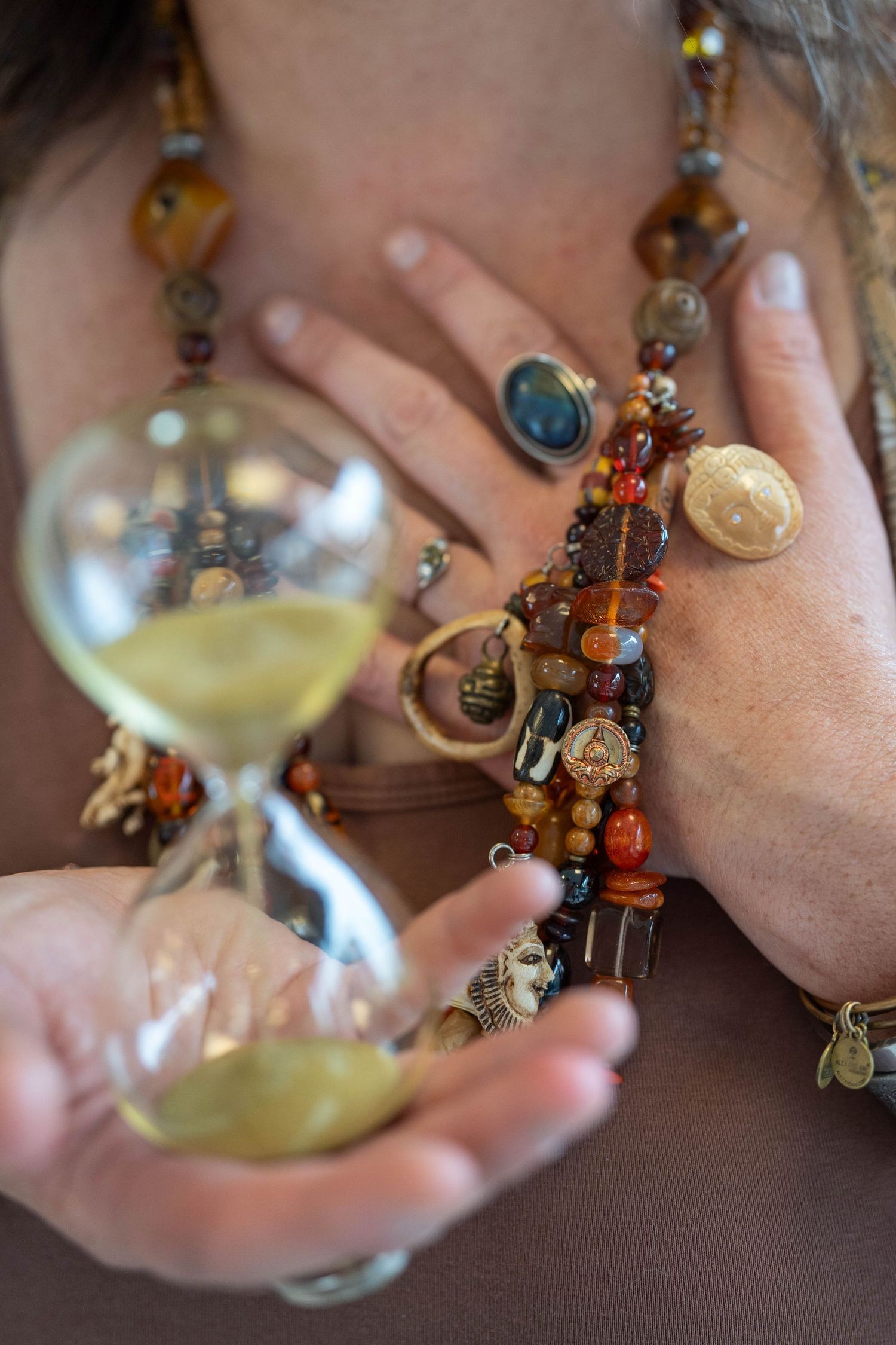 Woman holding hourglass with beaded bracelets, amber necklace, and gemstone rings, symbolizing time and spirituality.