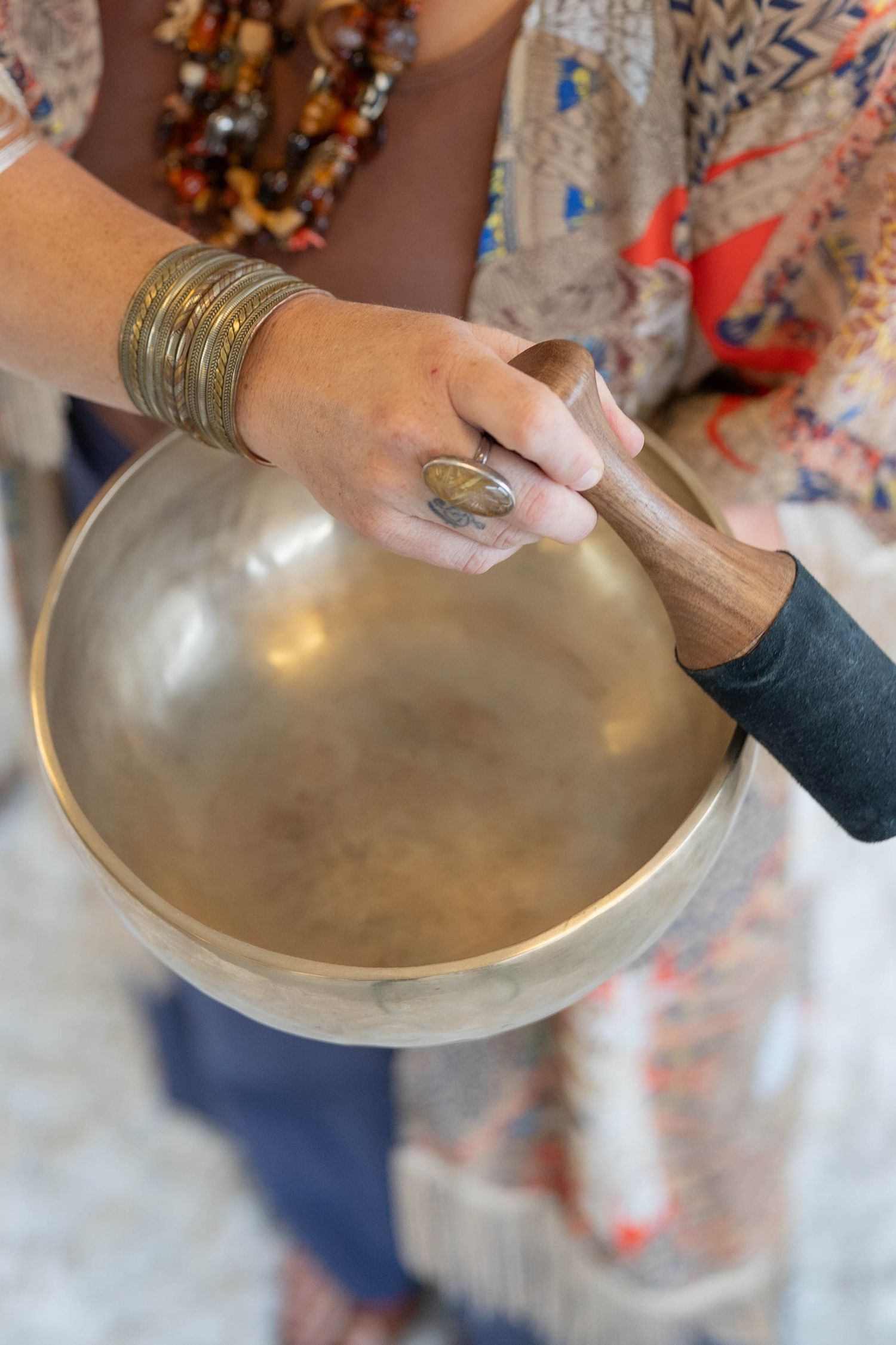 Woman playing a Tibetan singing bowl with a wooden mallet, wearing gold bangles and a large ring.