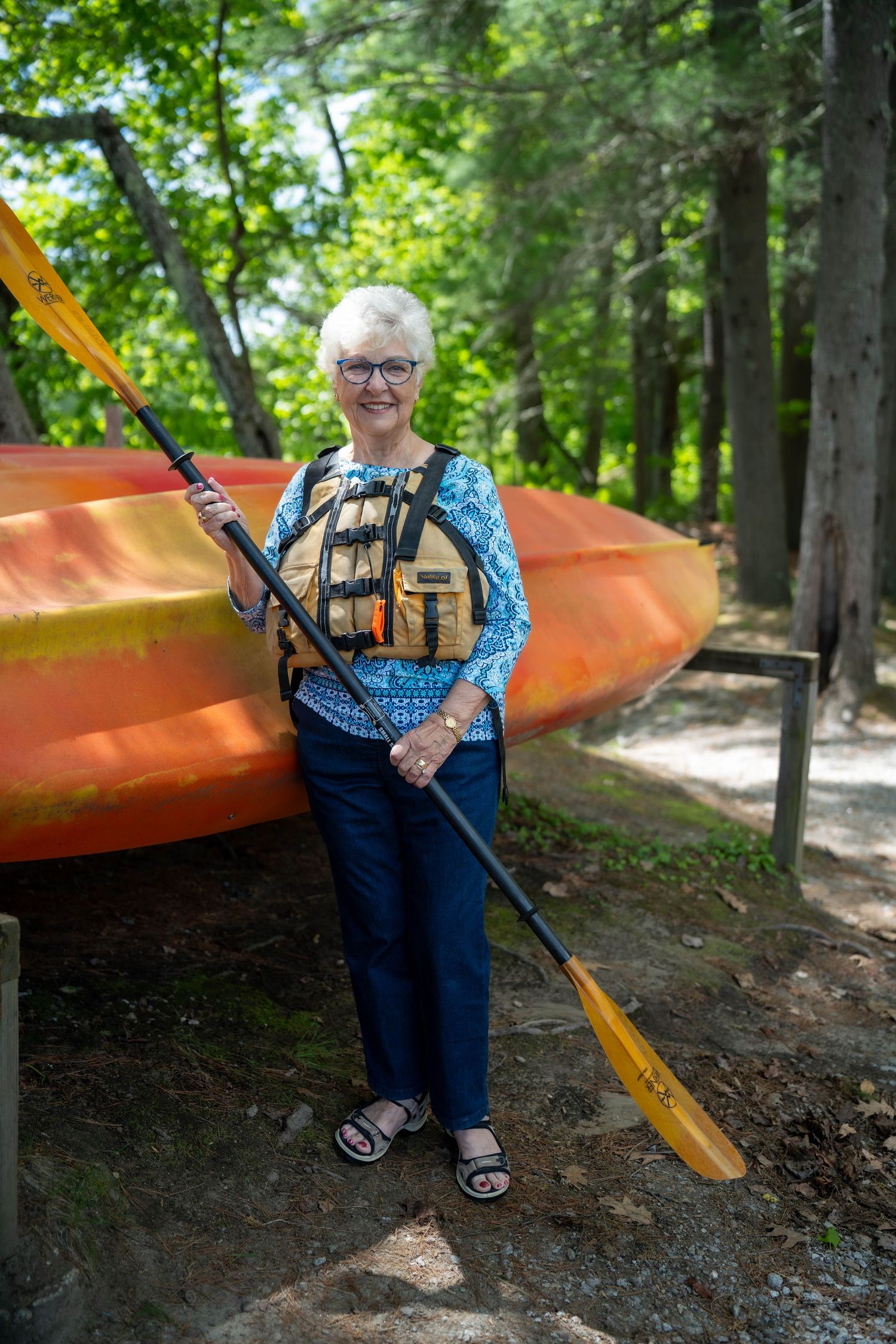 Smiling senior woman in life vest holds kayak paddle beside orange kayak in wooded area.