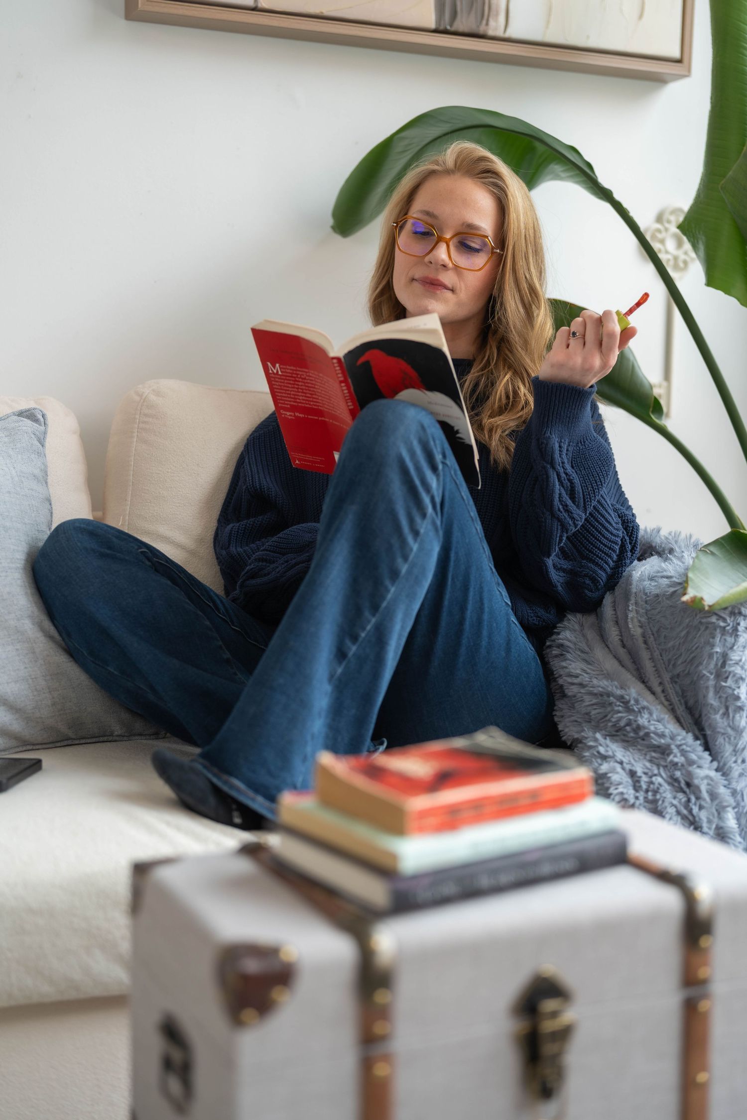Woman in glasses reading a red book on a cozy sofa with stacked books on a vintage trunk nearby.