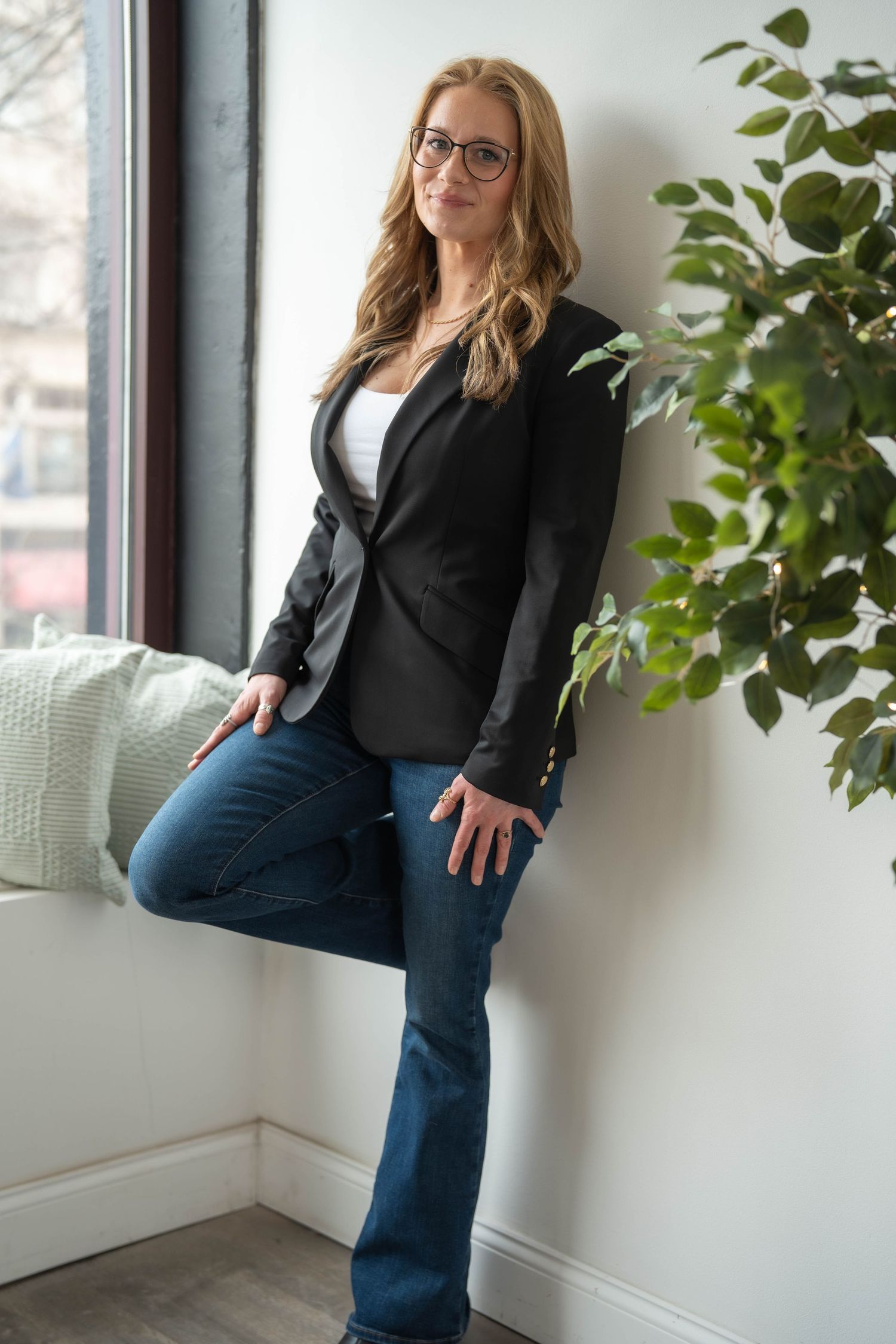 Woman in black blazer and jeans leaning against white wall near green plant and window.