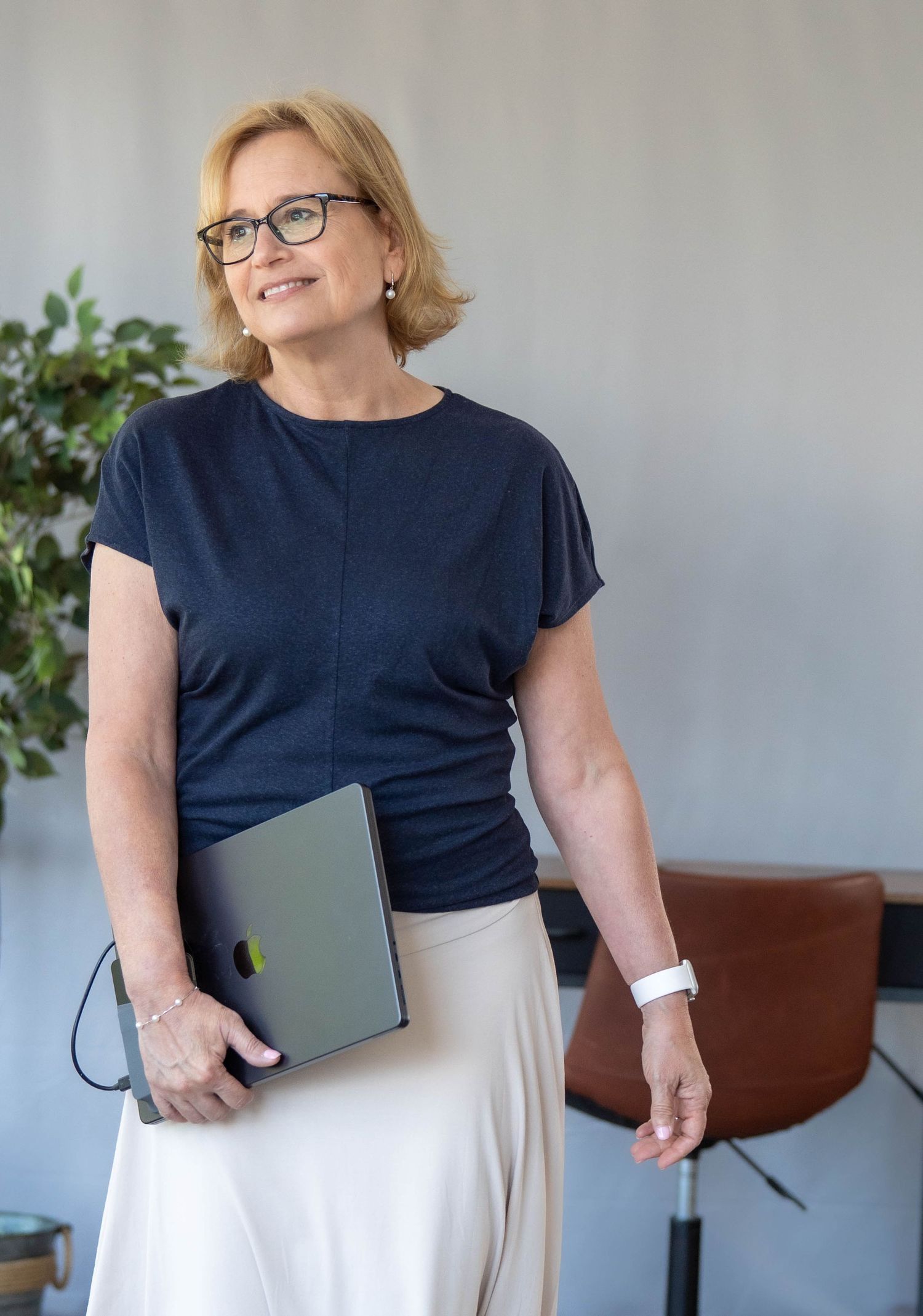 Professional woman in glasses holding a laptop, wearing navy top and beige skirt, smiling confidently in a modern office.