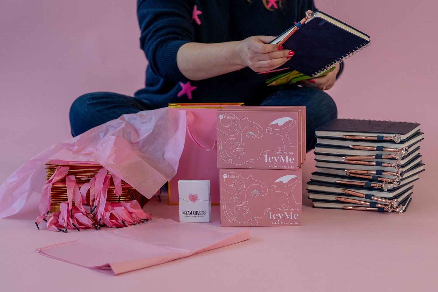 Person sitting cross-legged reading a journal surrounded by pink TeyMe boxes, flowers, and stacked notebooks on a pink background.
