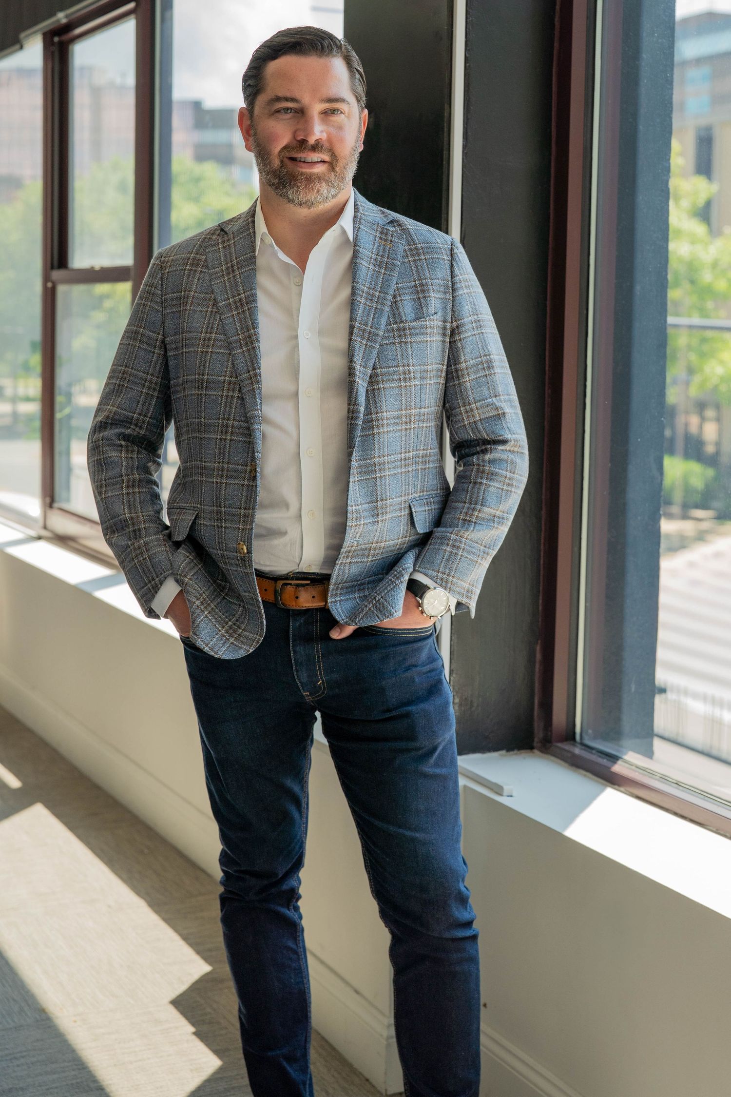 Professional man in plaid blazer and jeans poses confidently by large windows in a modern office building.