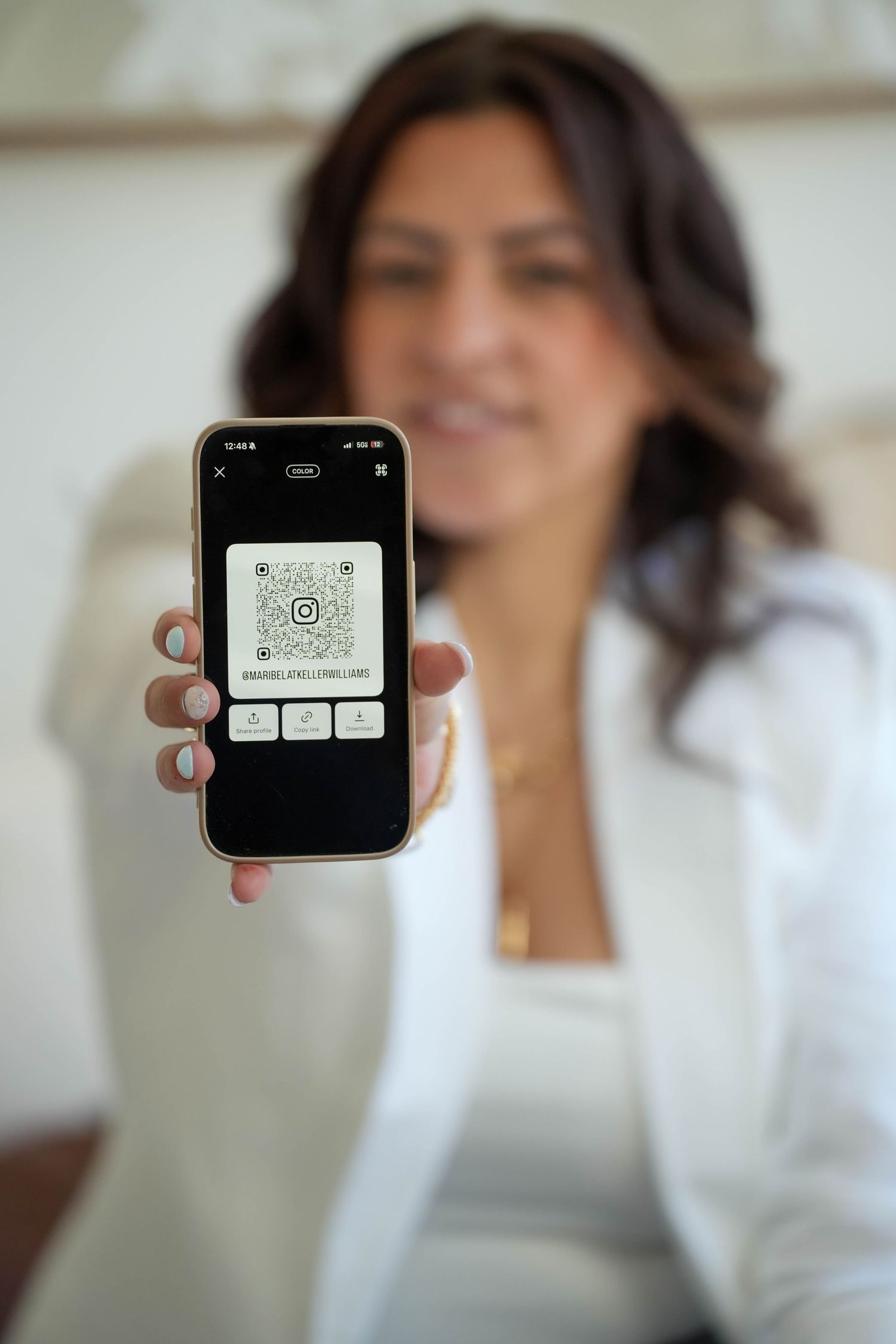 Woman in white blazer holding smartphone displaying an Instagram QR code toward the camera.