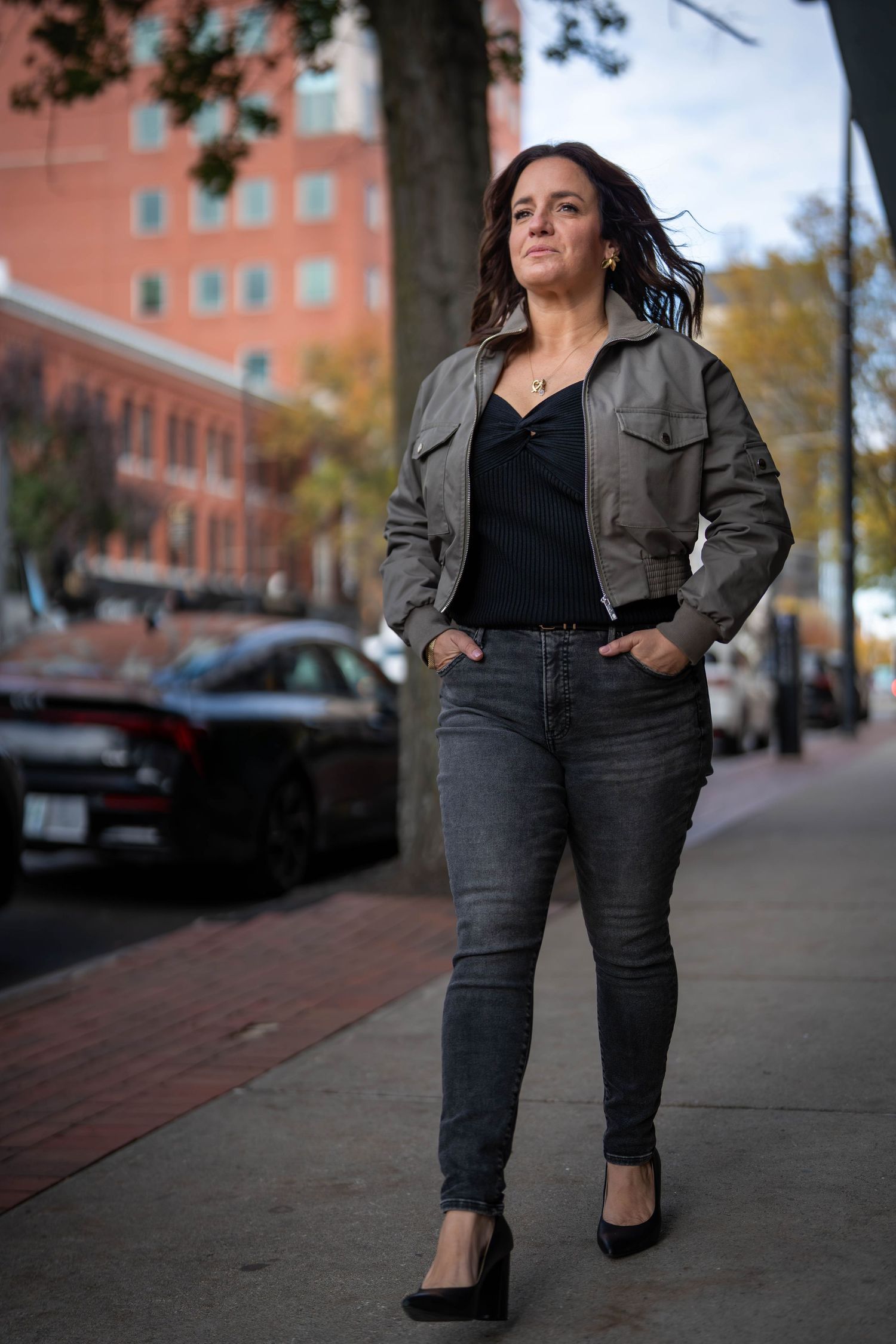 Woman in gray jacket and jeans poses confidently on urban sidewalk with brick buildings in background.