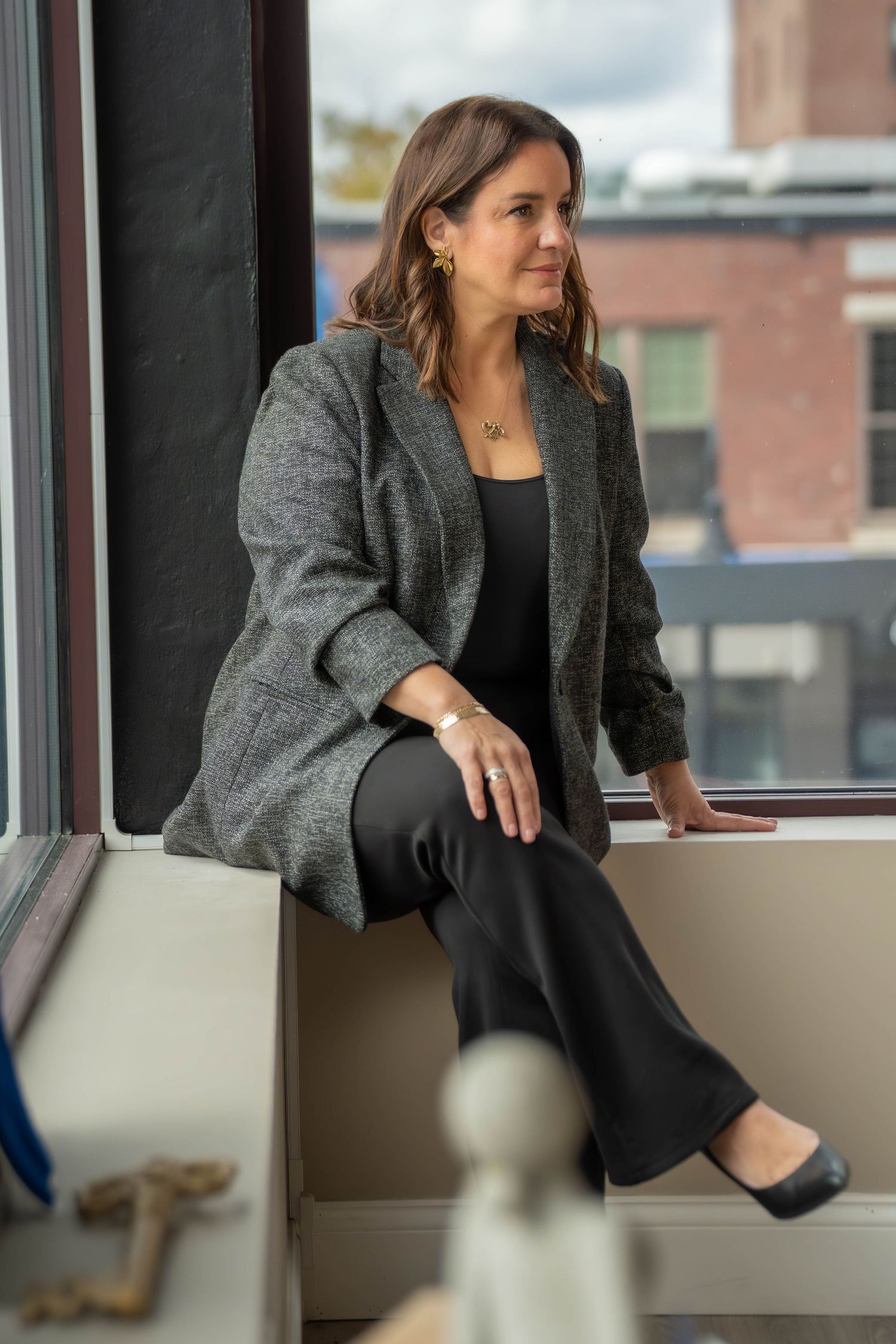 Woman in gray blazer and black pants sits on windowsill, gazing thoughtfully with urban backdrop.