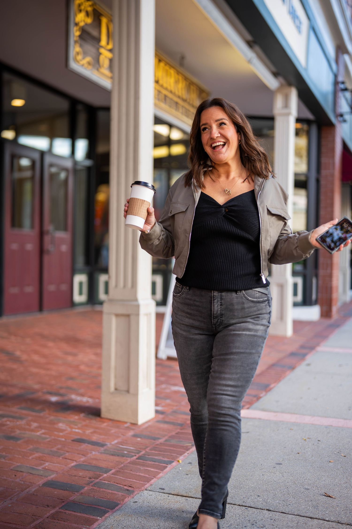 Smiling woman walking on city sidewalk holding coffee cup and phone, wearing gray jacket and black jeans.