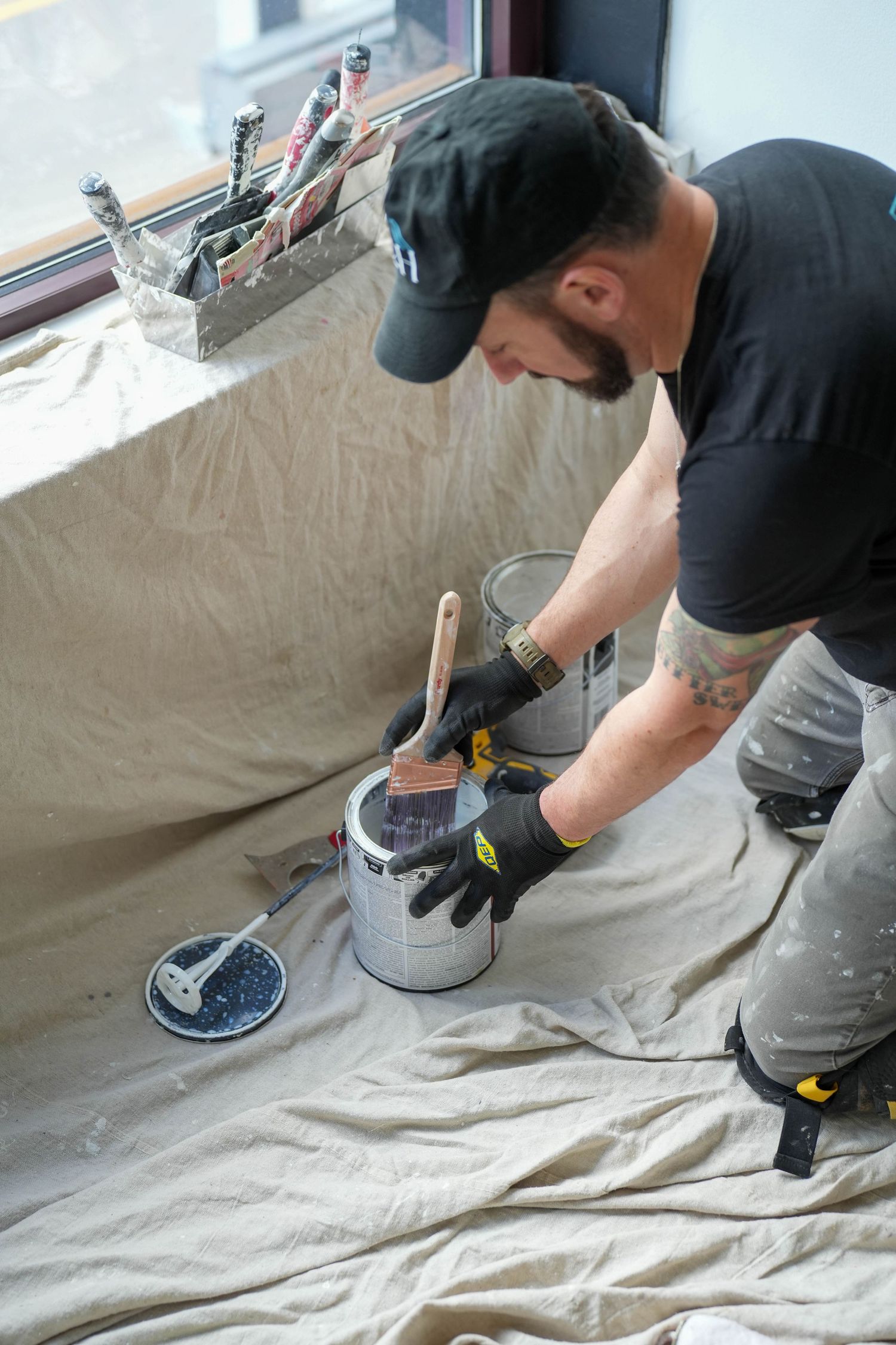 A painter in black gloves dips a brush into a paint can on a drop cloth near a window.