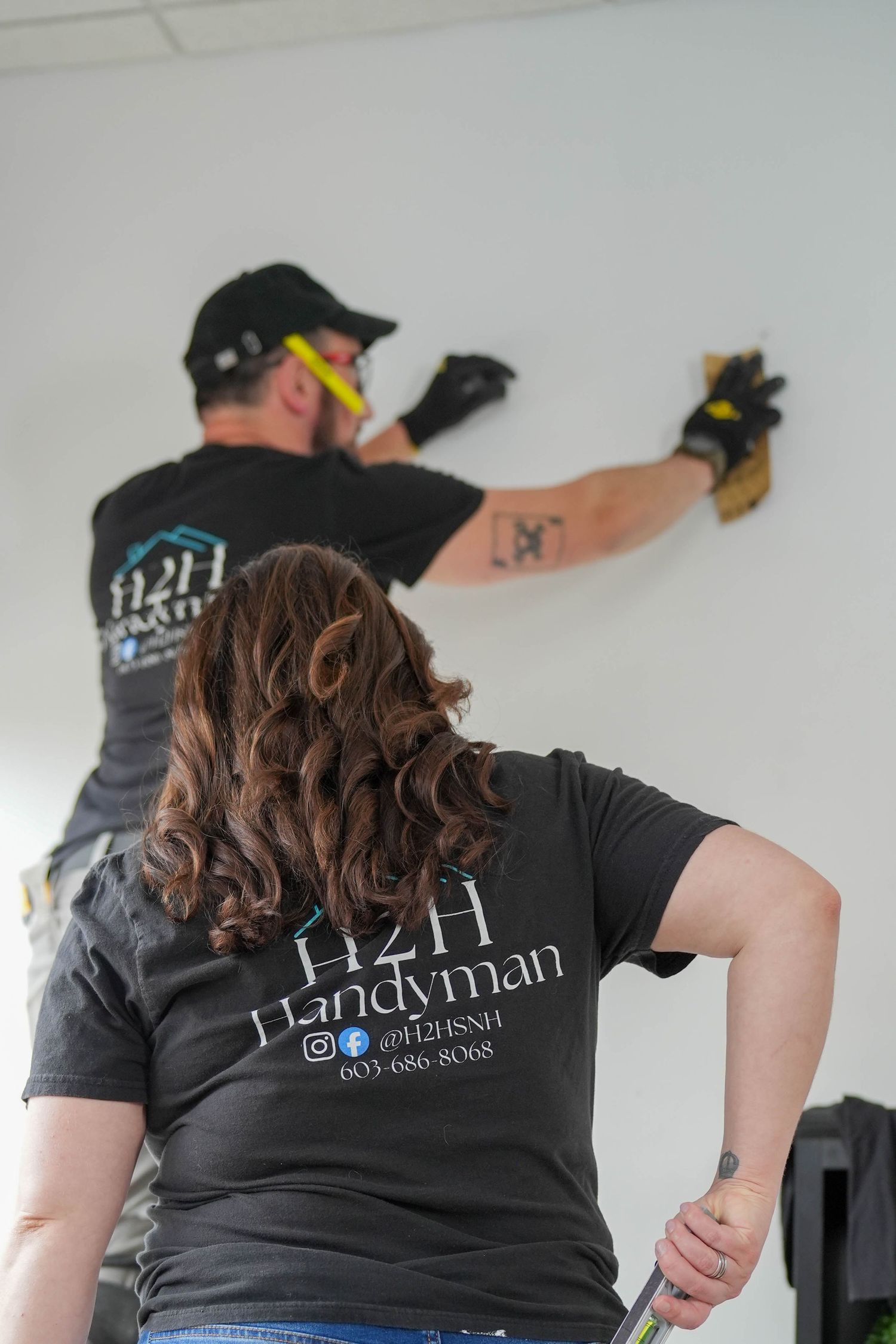 Two H2H Handyman workers in branded black t-shirts performing wall repairs with tools in a bright interior space.