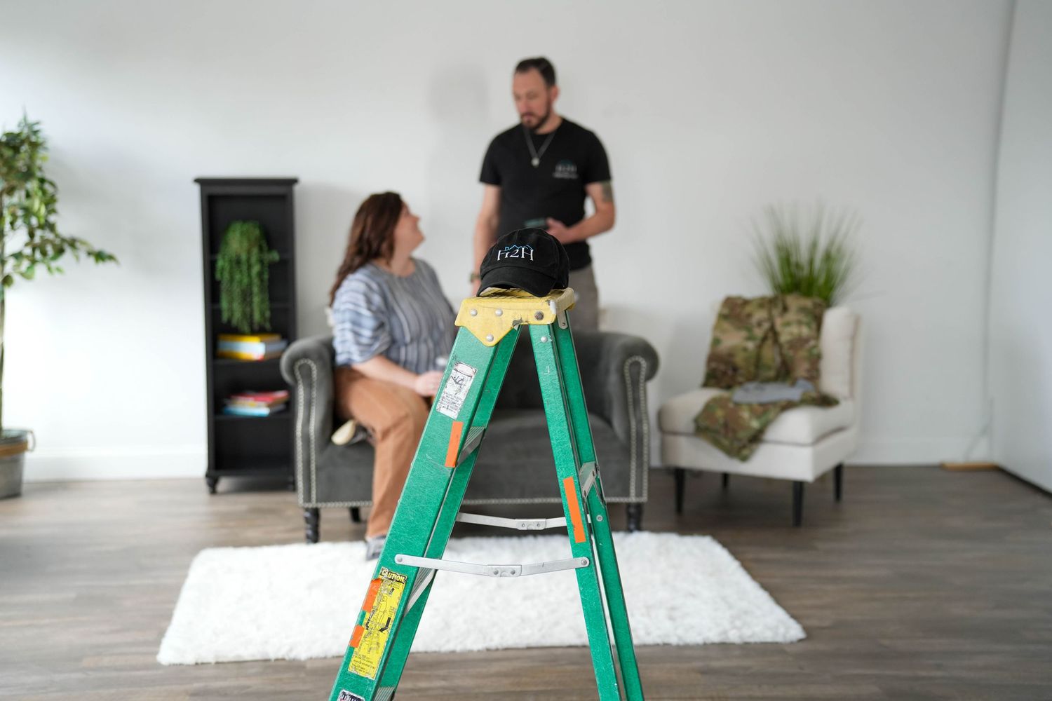 Woman sitting on a green ladder while a man stands nearby in a bright modern living room.