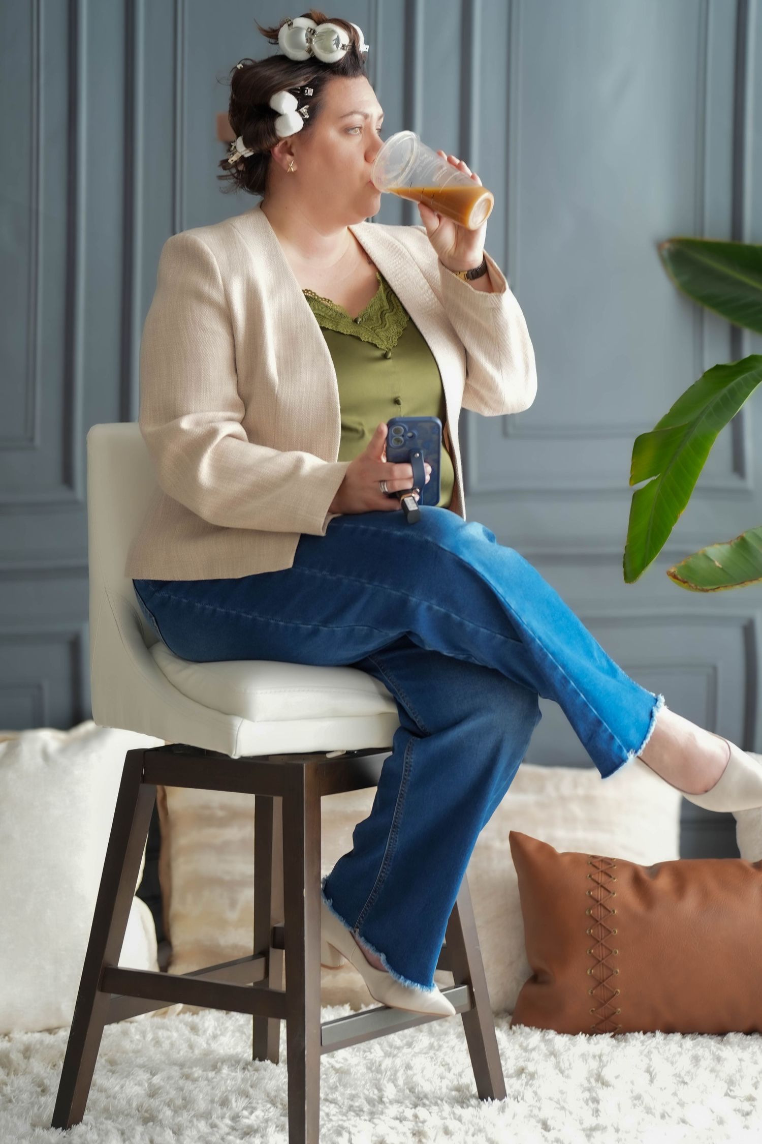 Woman with hair rollers sitting on barstool drinking juice while holding phone in stylish outfit.