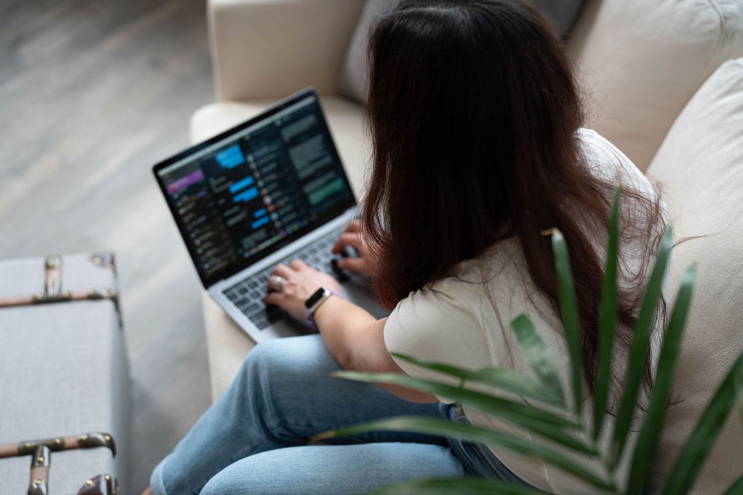 Woman sitting on couch using laptop with dark coding interface, working from home in casual jeans and white top.