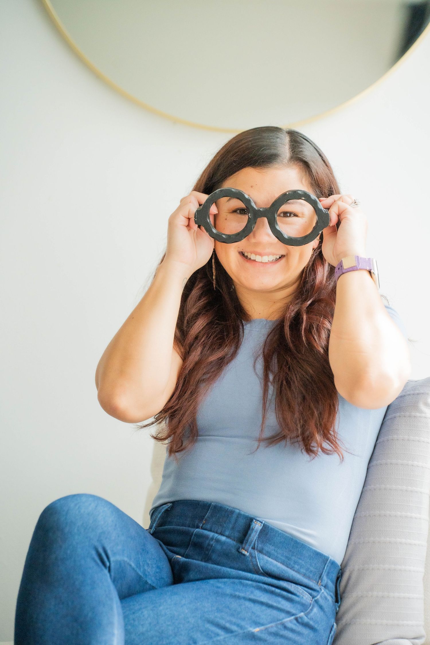 Woman smiling while holding oversized novelty glasses in front of her eyes, sitting casually in jeans and a blue top.