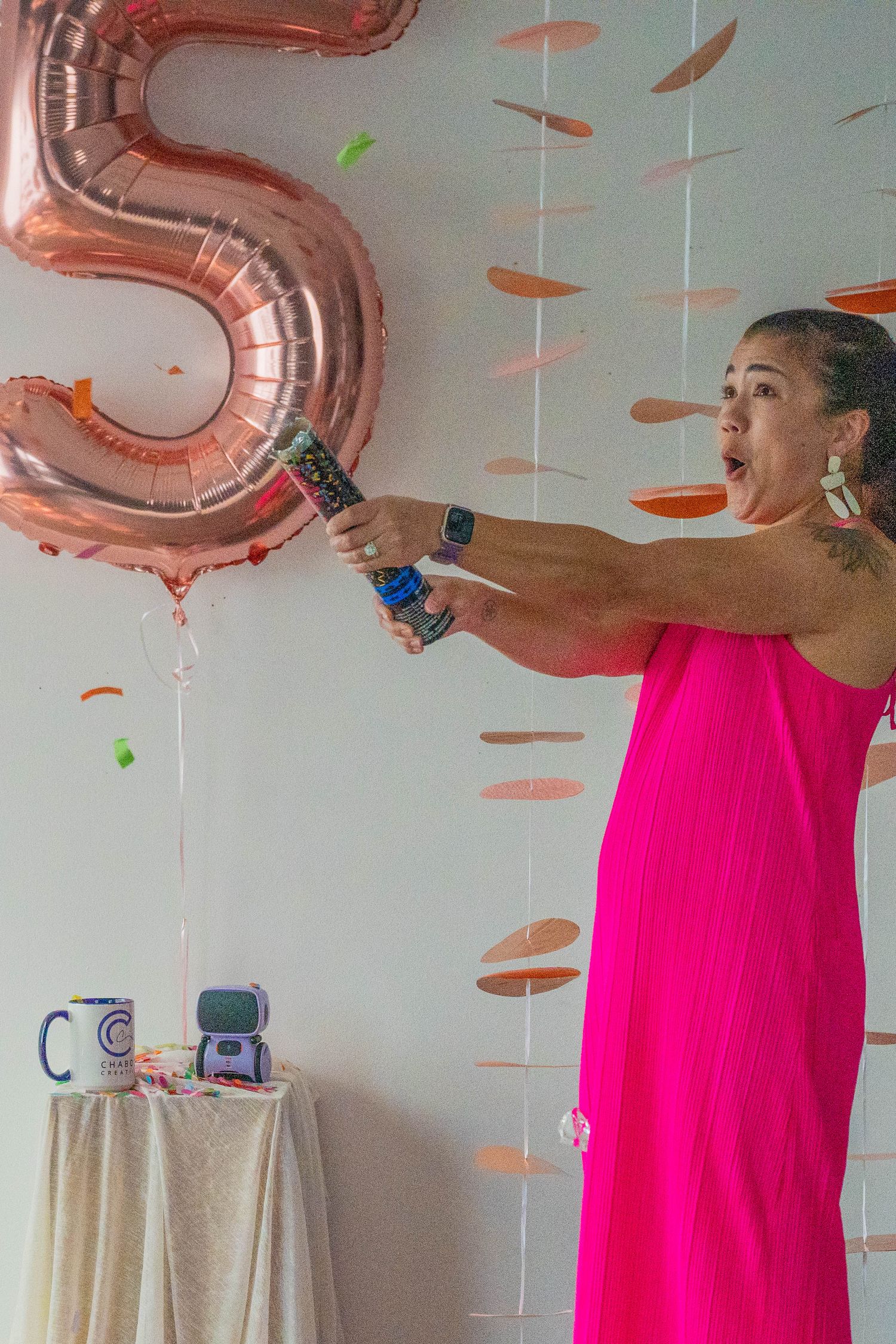 Woman in pink dress popping confetti cannon near rose gold number 5 balloon at a birthday party celebration.