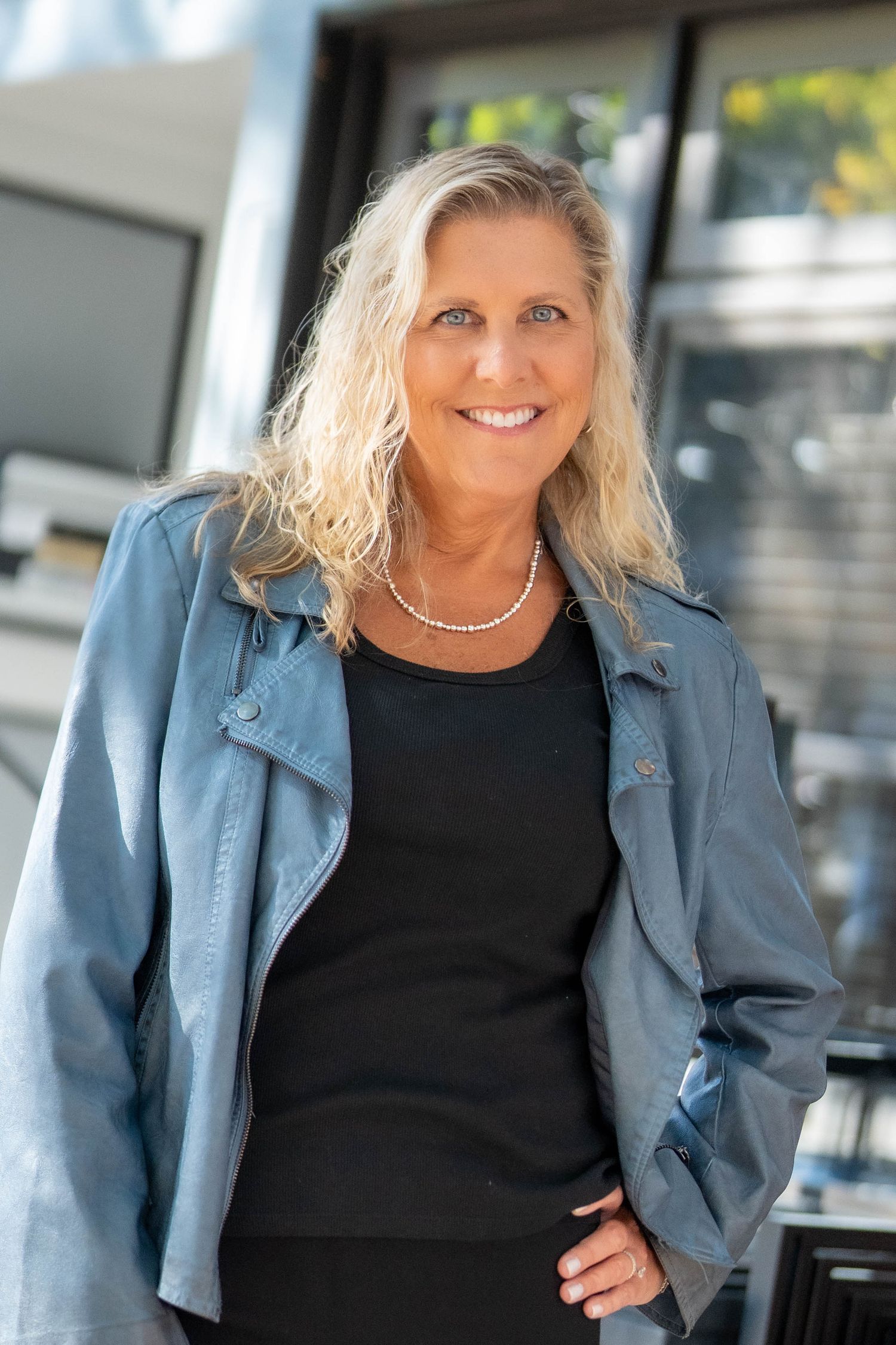 Smiling blonde woman in blue leather jacket and black top with pearl necklace posing outdoors.