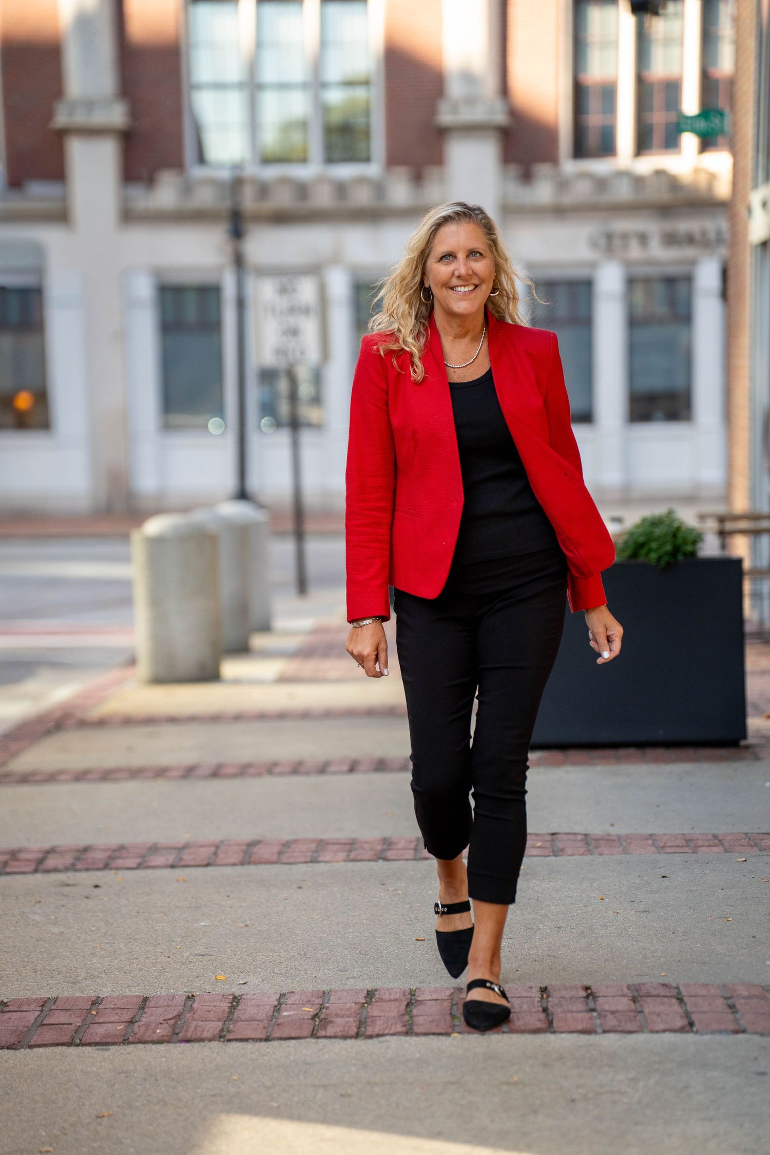 Woman in red blazer and black outfit walking confidently on a brick plaza near City Hall.