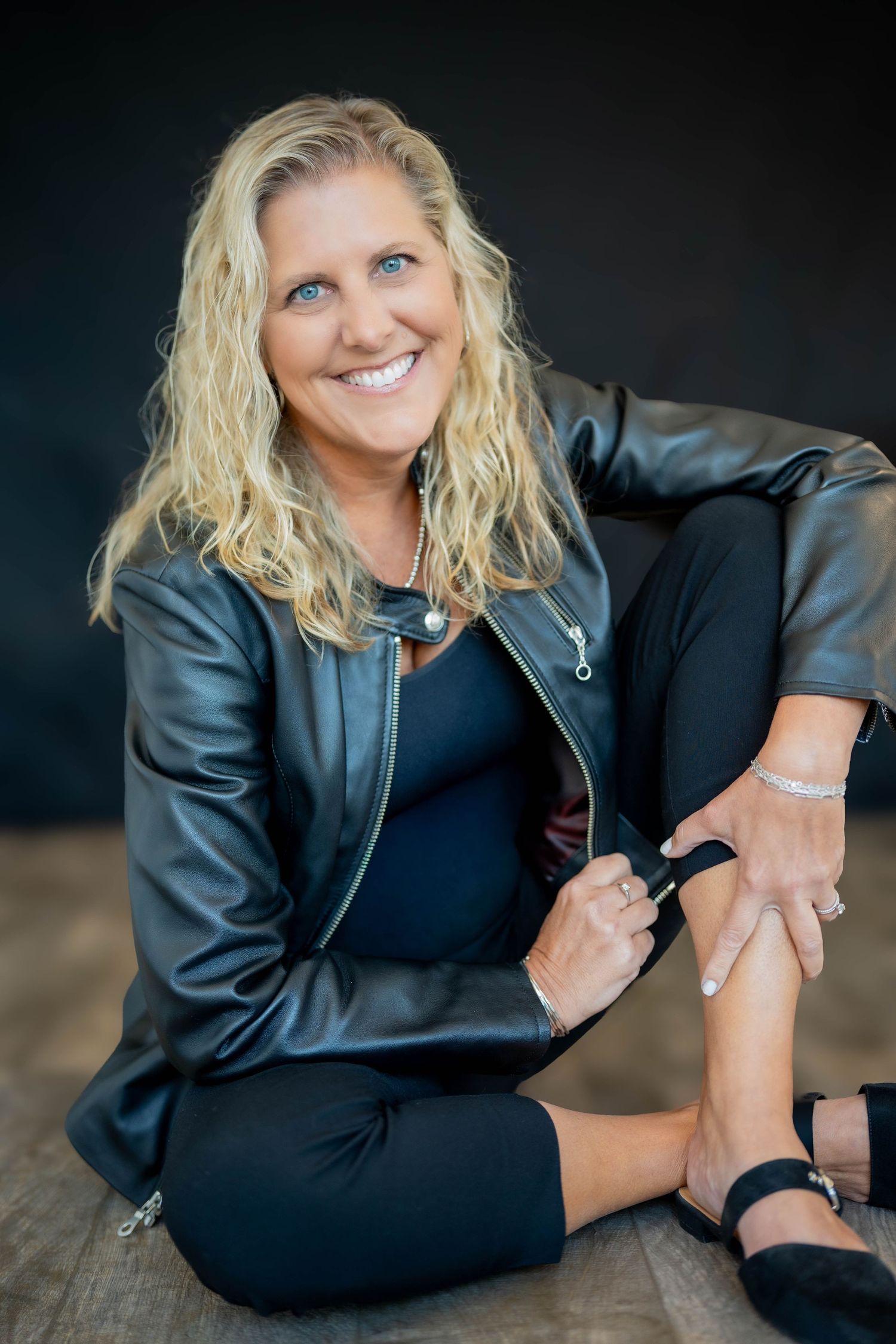 Smiling blonde woman in black leather jacket and pants sitting casually on floor against dark background.