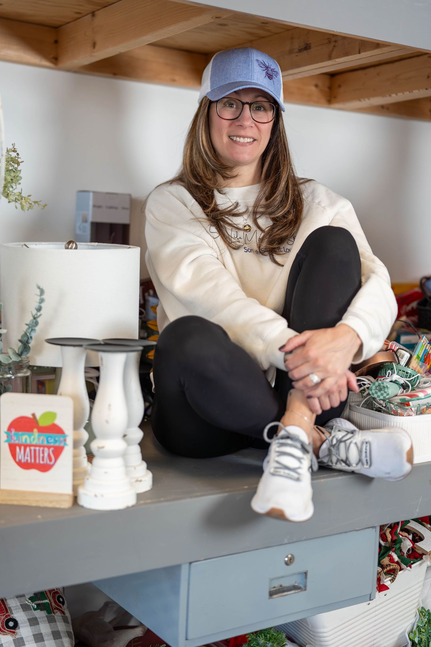 Woman in blue cap and white sweatshirt sits smiling on a gray shelf surrounded by home decor items.