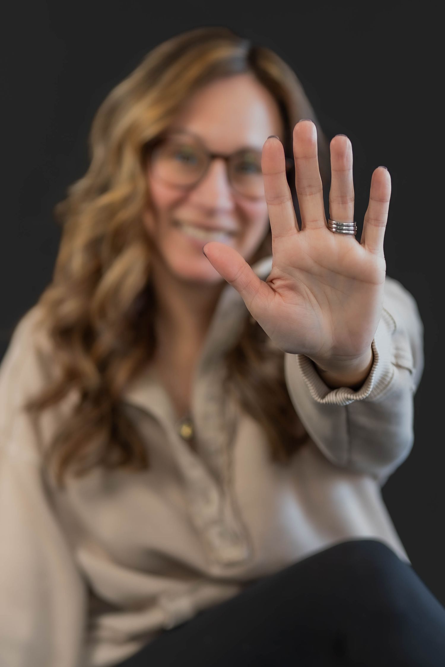 Smiling woman with glasses and wavy hair holds her palm up toward the camera, wearing a ring, against a dark background.