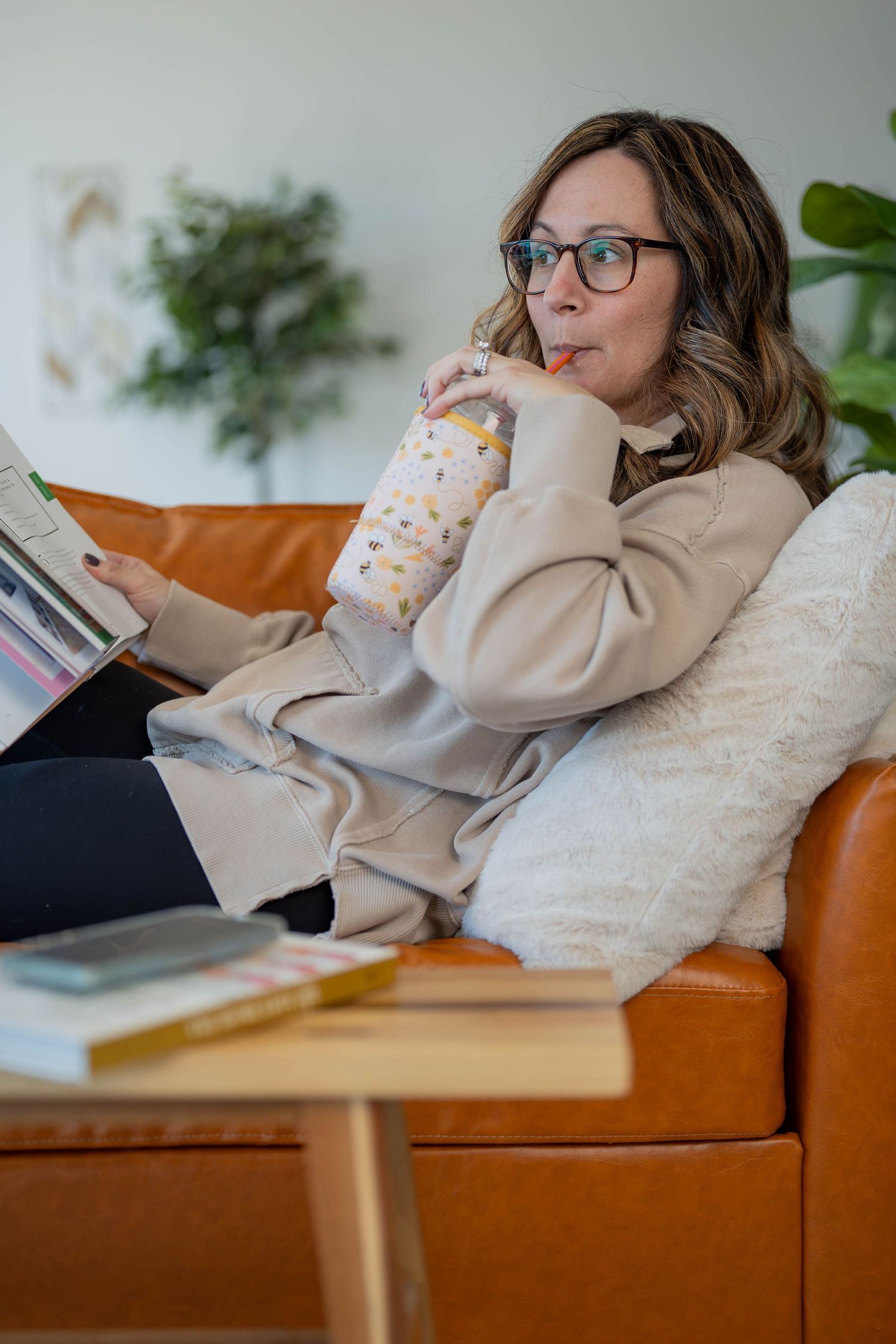 Woman in glasses relaxing on leather sofa, sipping from tumbler while reading a book, with plants in background.