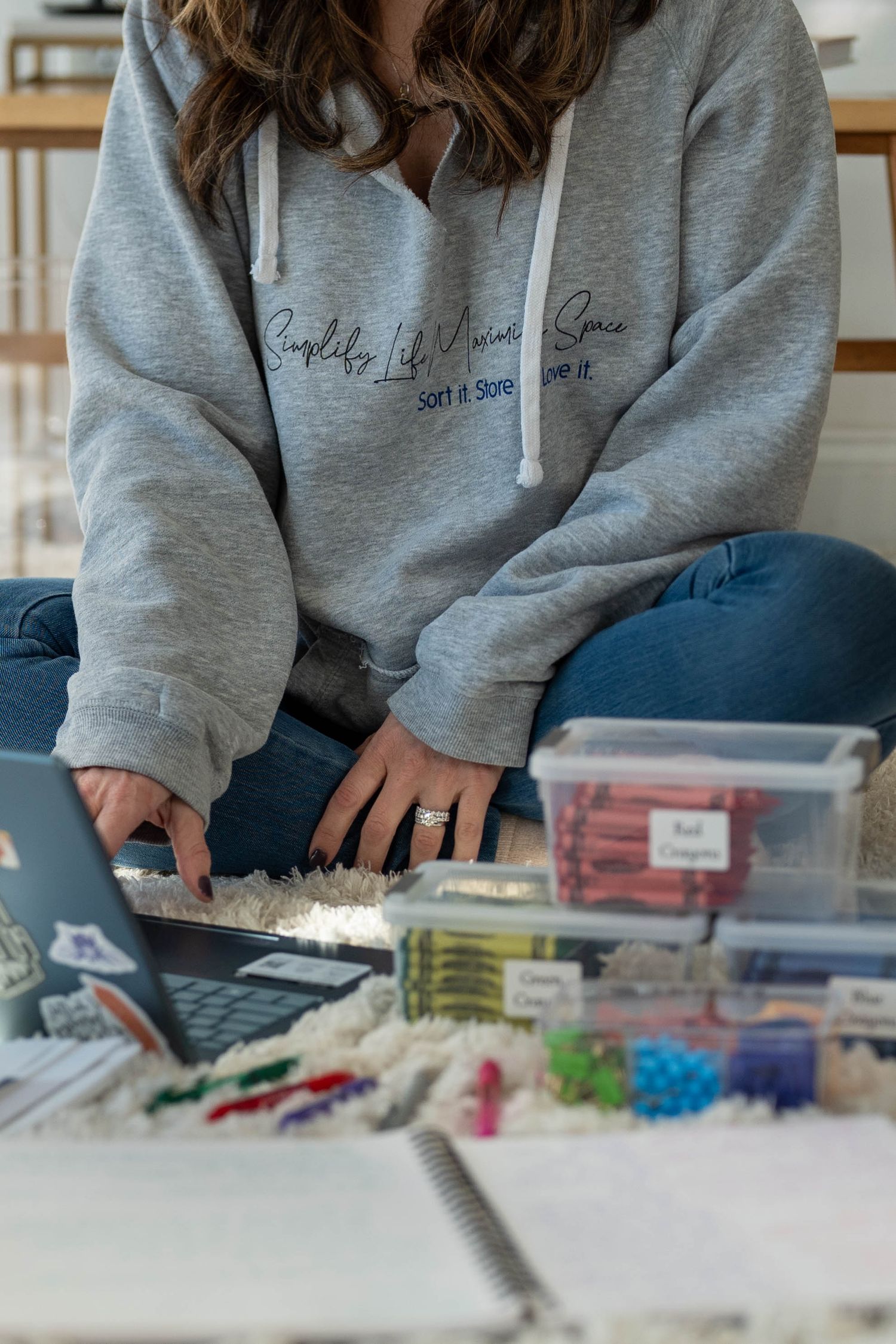 Woman in gray hoodie organizing supplies, typing on laptop surrounded by labeled storage containers and craft materials.