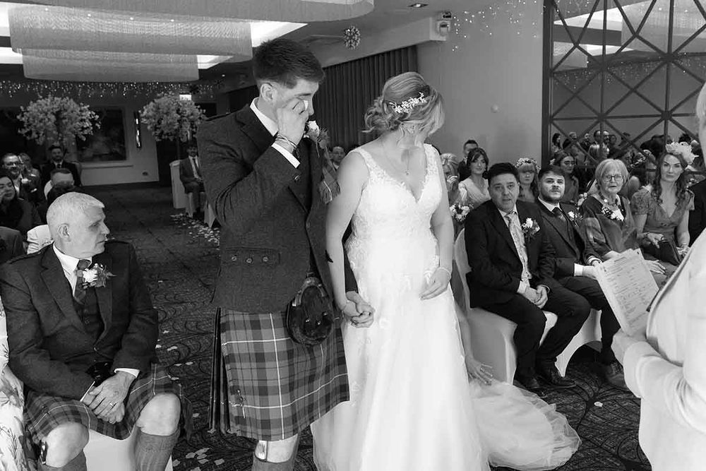 Black and white wedding photo of emotional groom in kilt wiping tears beside bride in white gown during ceremony.